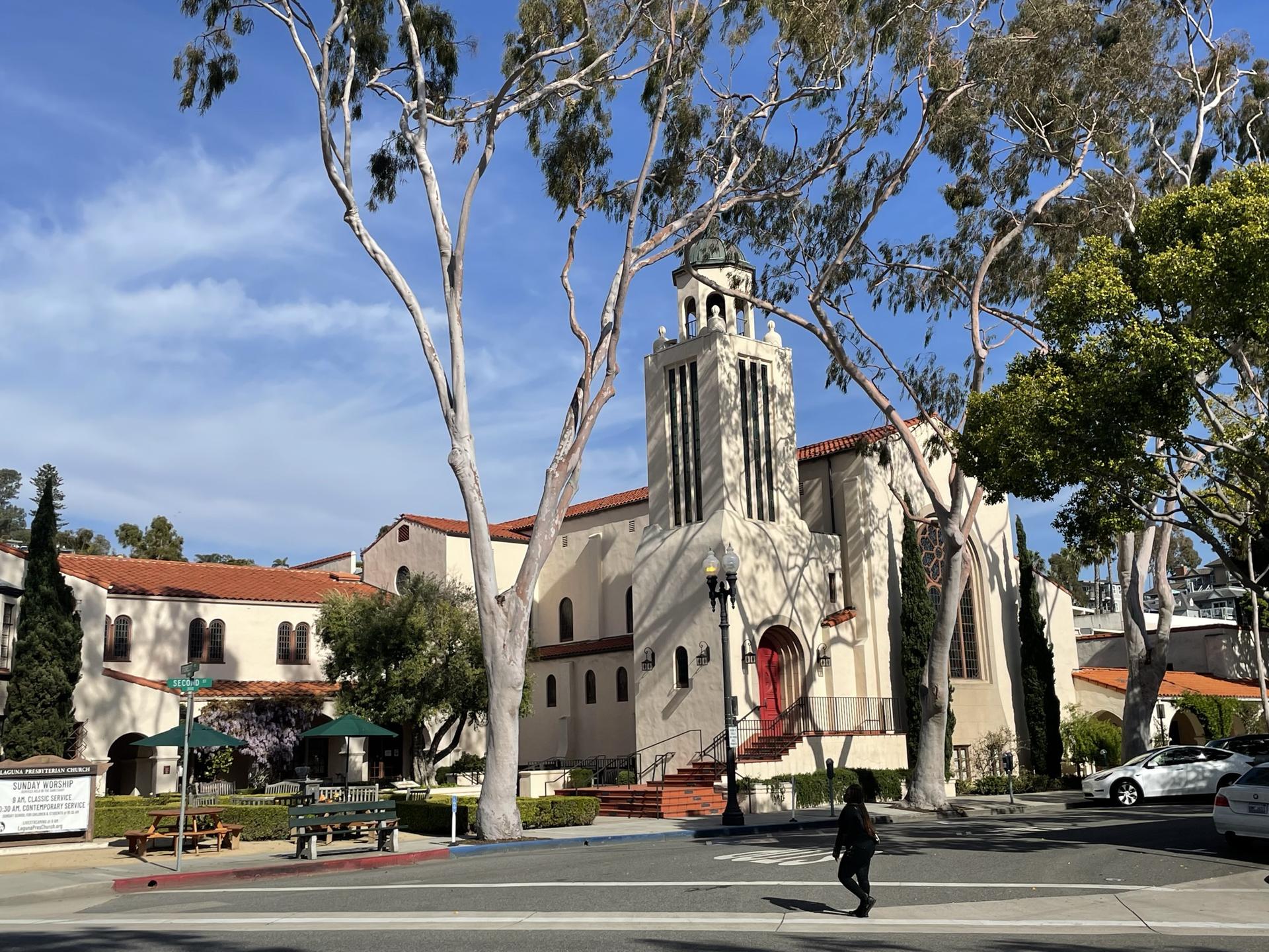 A person walks past a large building with a bell tower on a sunny day.
