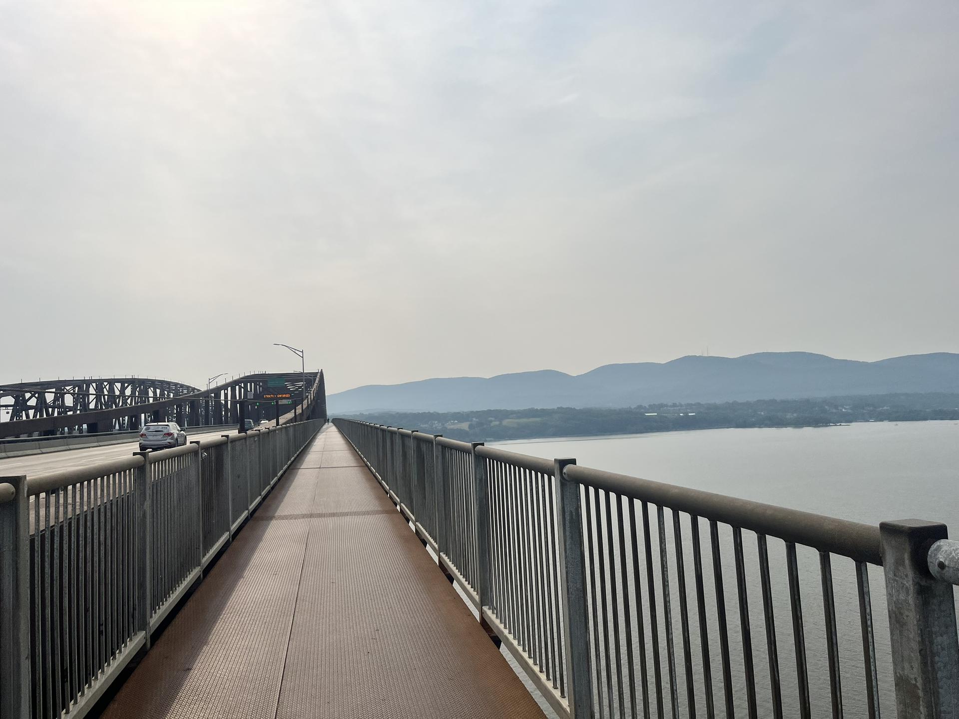 Long bridge with pedestrian walkway and vehicle lanes over water, hazy sky.