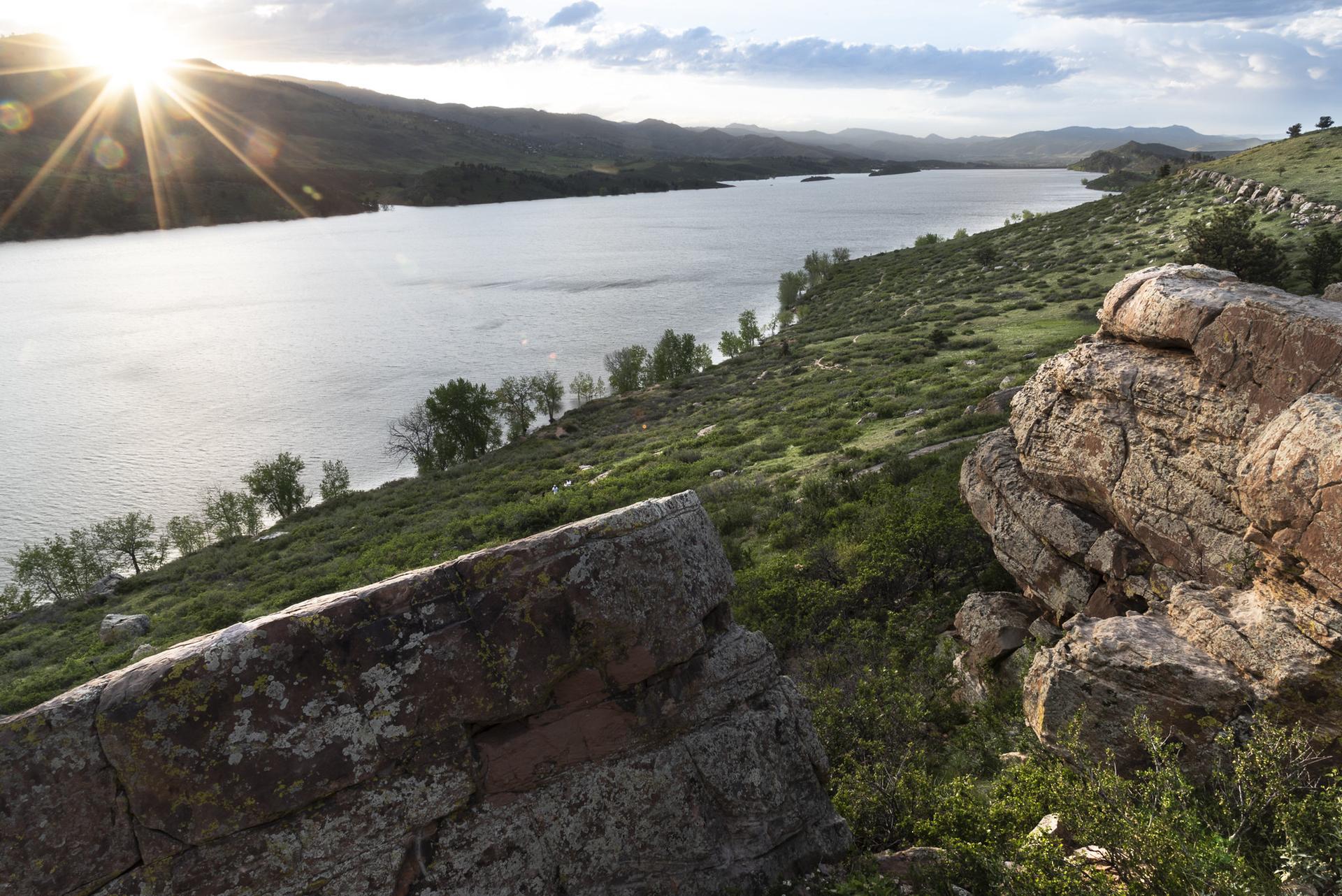 Large rocks on a green hillside overlooking a lake with a bright sunburst.