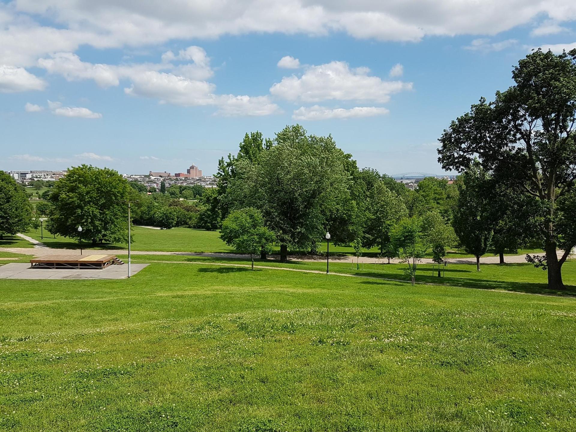 A wide, sunny green park with trees, a stage, and distant city views.