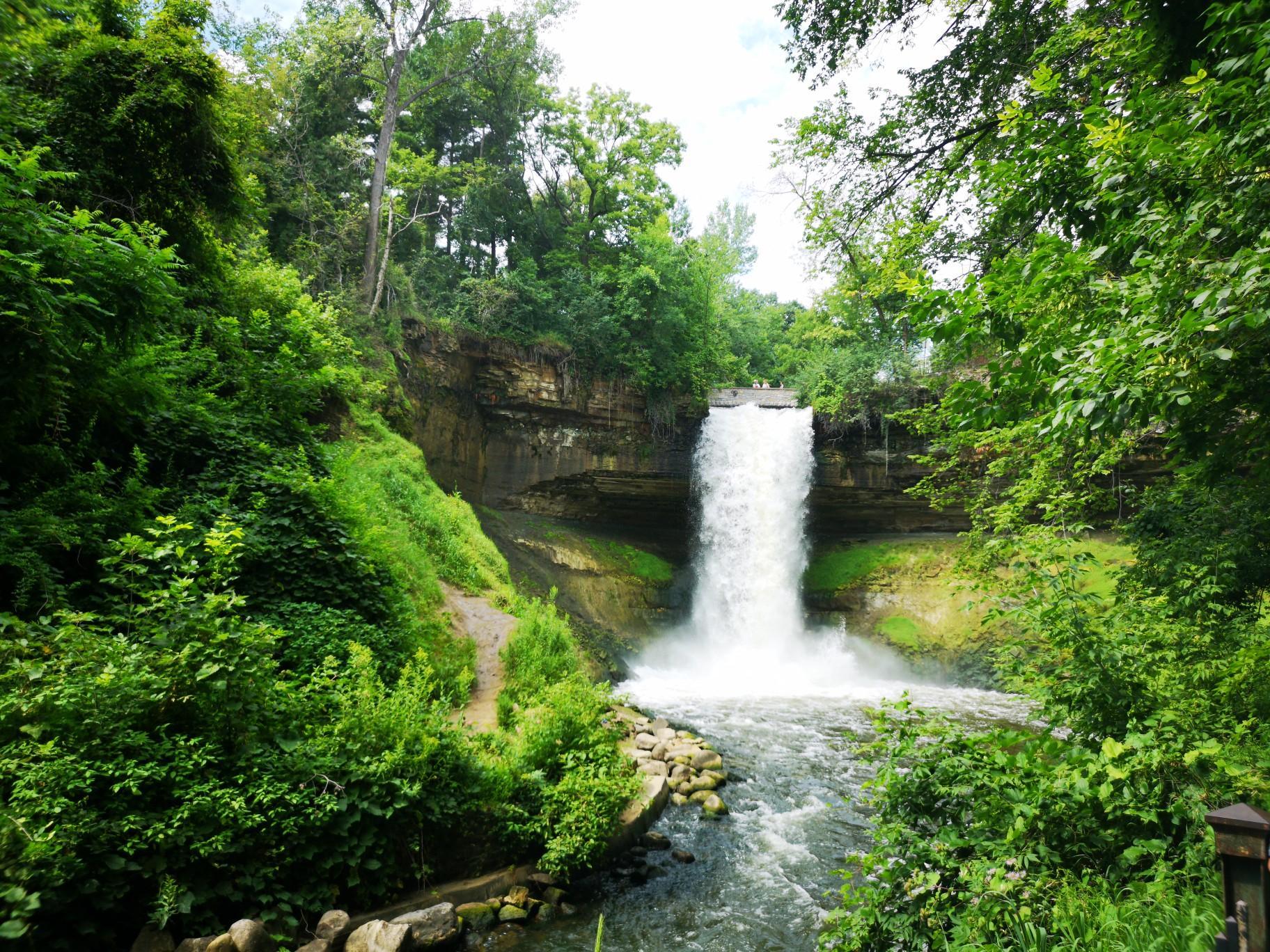 Wide view of a powerful waterfall surrounded by dense green forest. Sunny day.