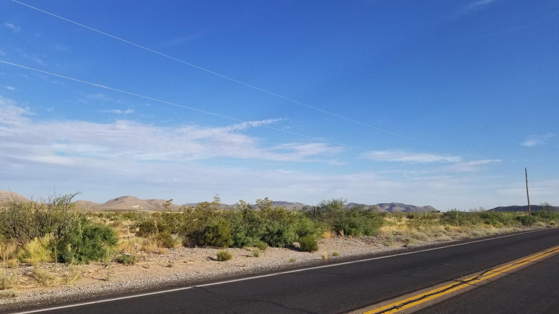 Desert road with scrub brush, distant mountains, and power lines under a bright blue sky.