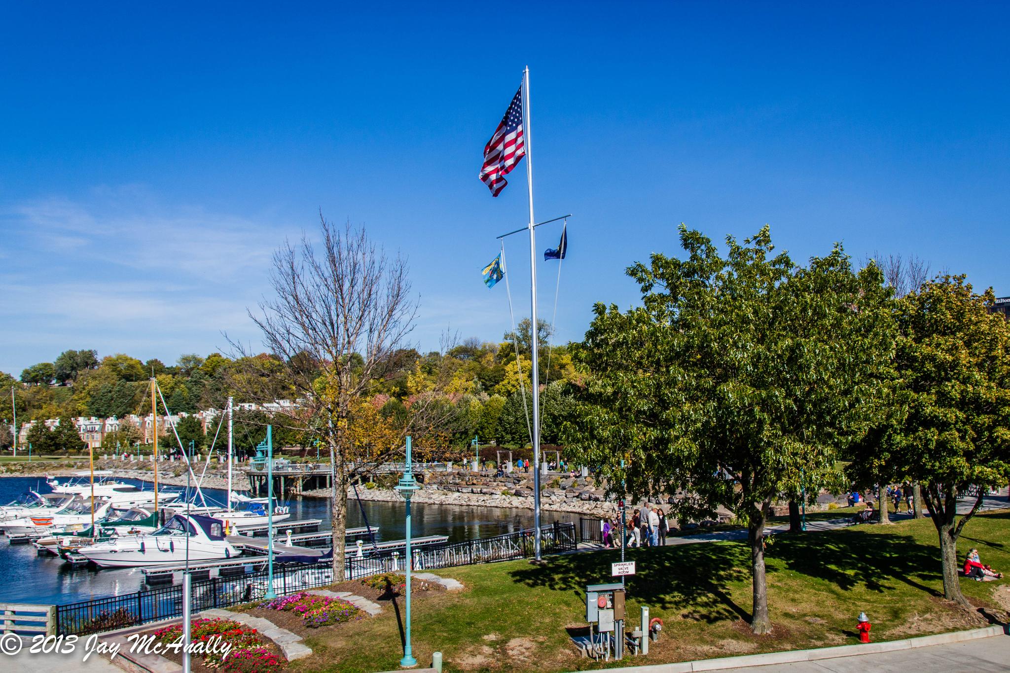 Marina with boats, flags flying, green trees, and distant people under a clear sky.