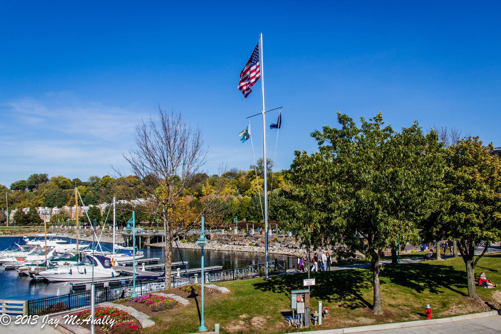 Marina with boats, flags flying, green trees, and distant people under a clear sky.