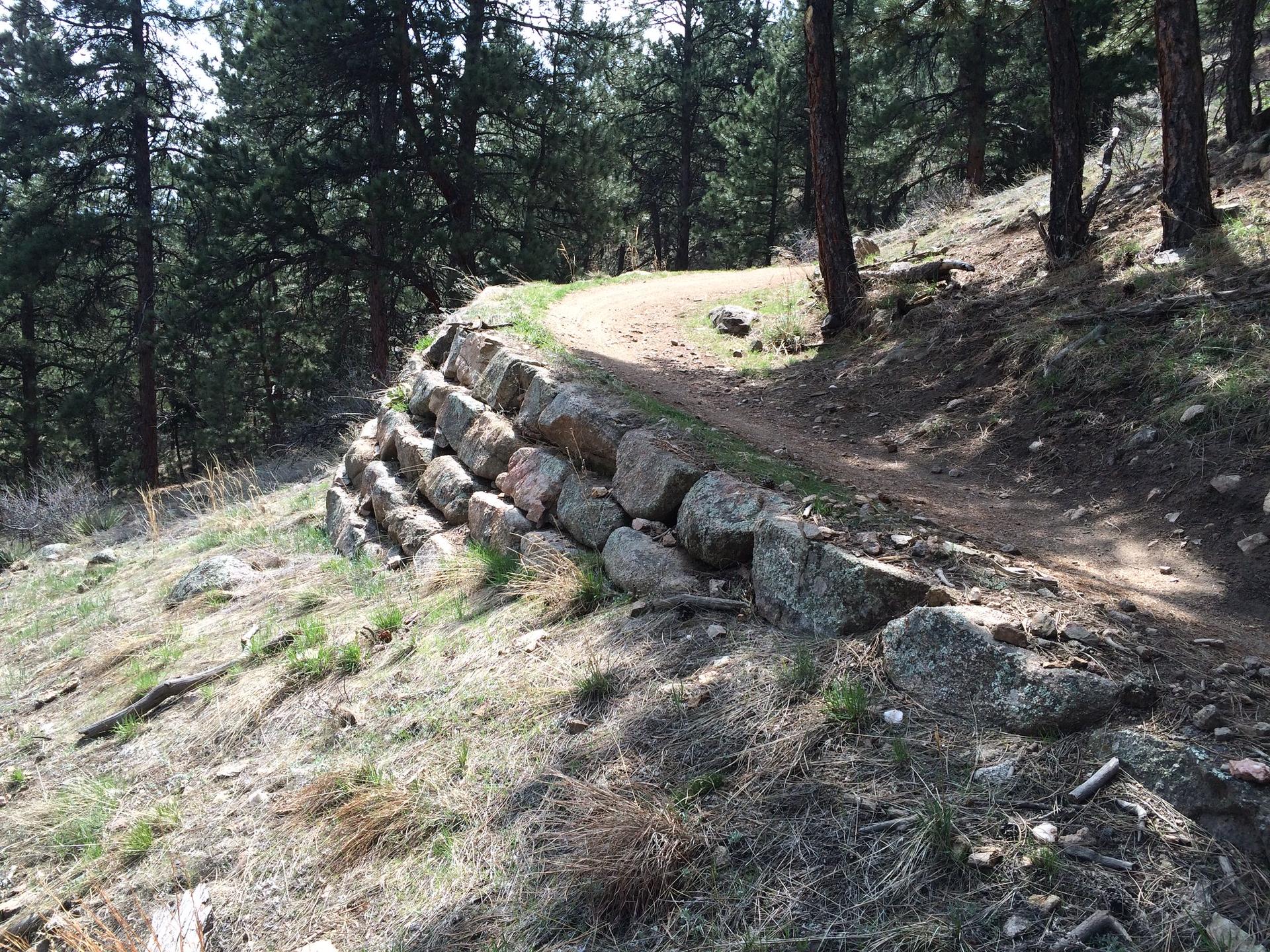 Dirt trail winding uphill next to a stacked rock retaining wall in a forest.