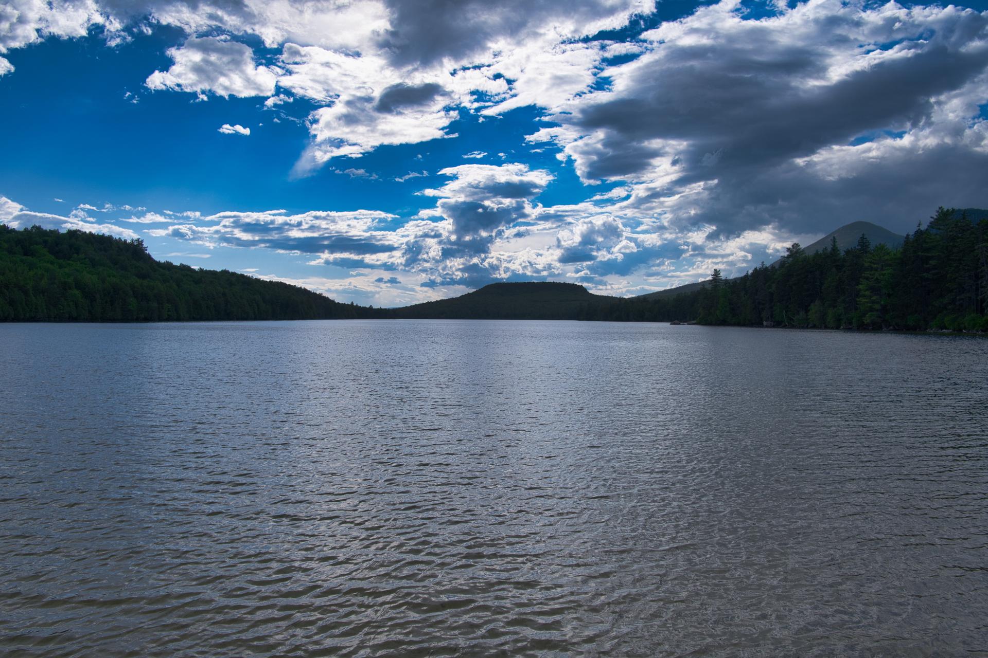 Vast lake surrounded by dark forested hills under a dramatic cloudy sky.