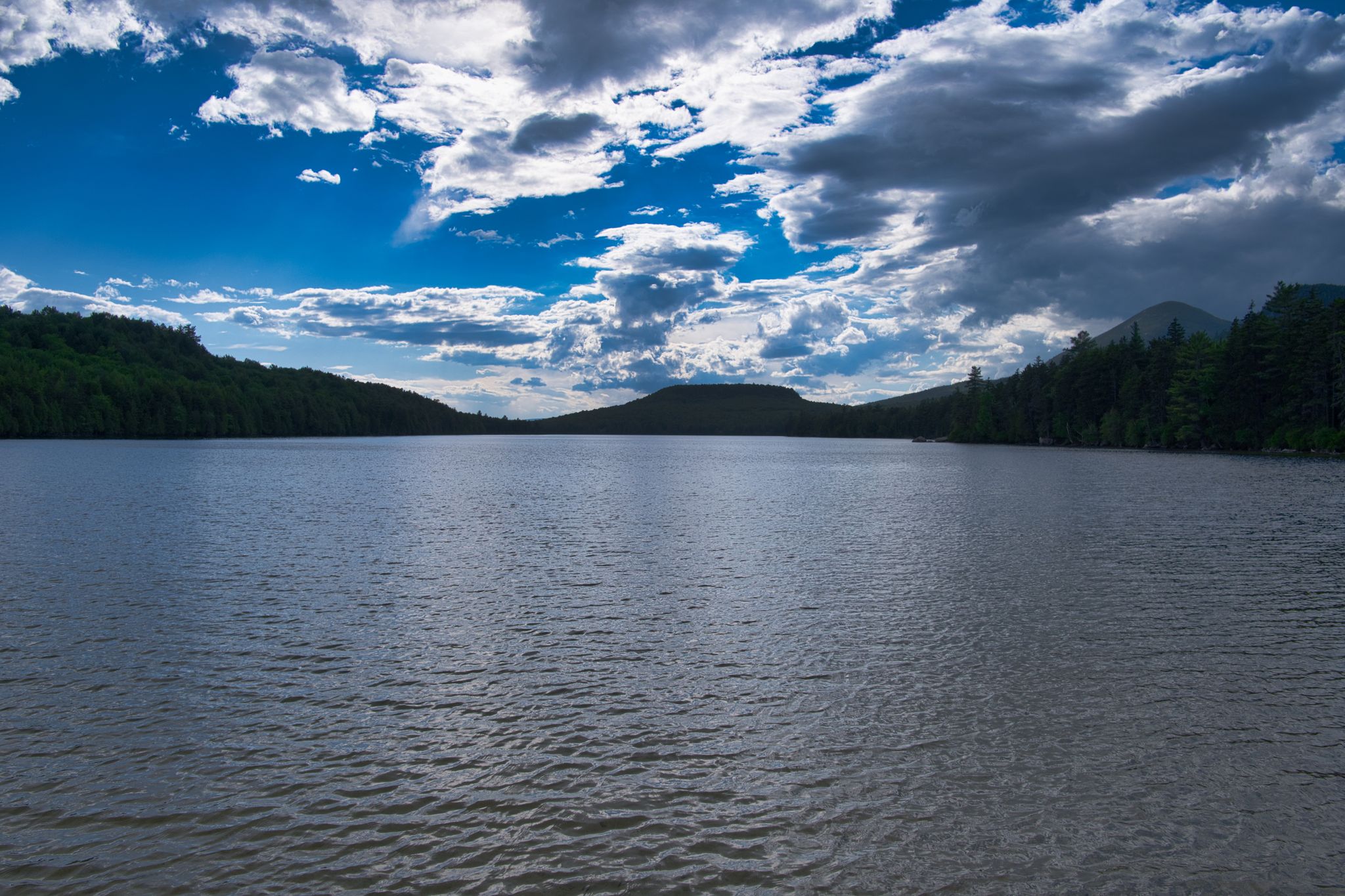 Vast lake surrounded by dark forested hills under a dramatic cloudy sky. - Christine Lake to South Pond