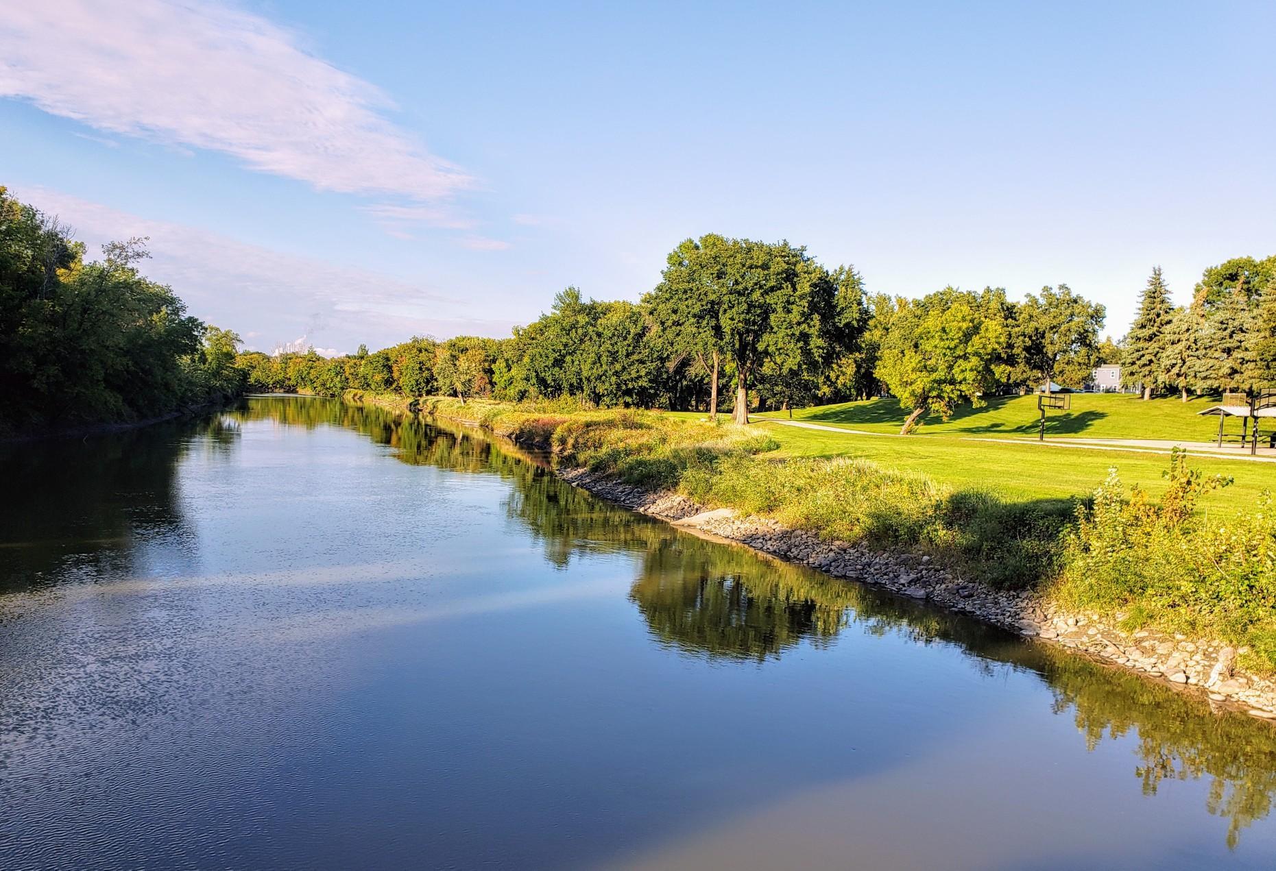 Wide river reflecting blue sky, flanked by lush green banks and trees.