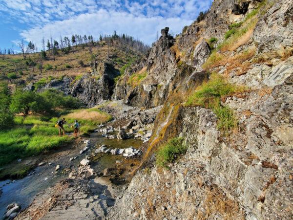 Two people by a rocky stream in a sunny, mountainous landscape with sparse trees.