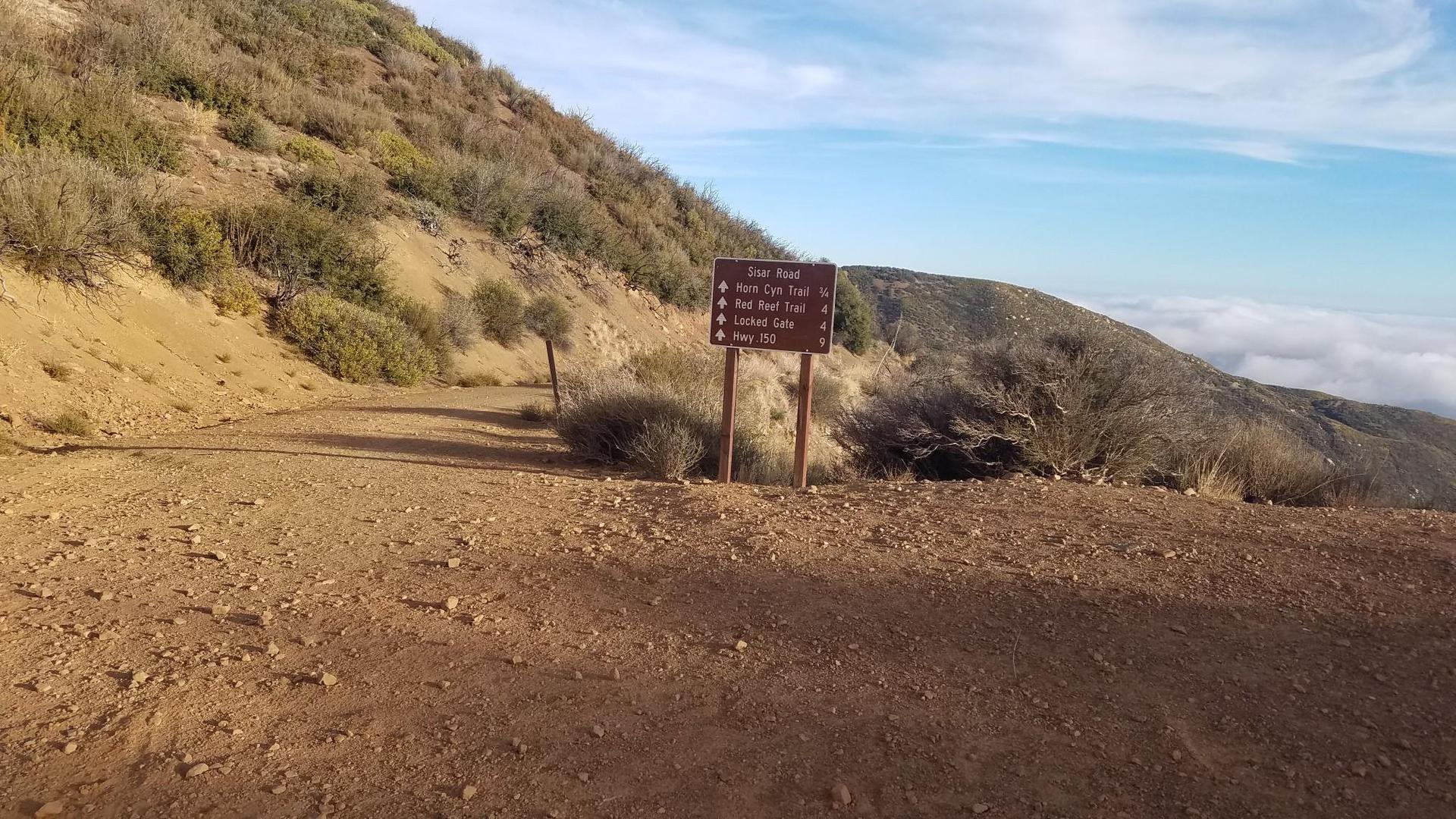 Wooden trail sign on a dirt path with a steep, sunny hillside and clouds below.