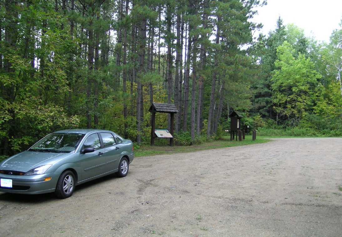 Green car parked at a dirt trailhead with wooden signs and tall trees.