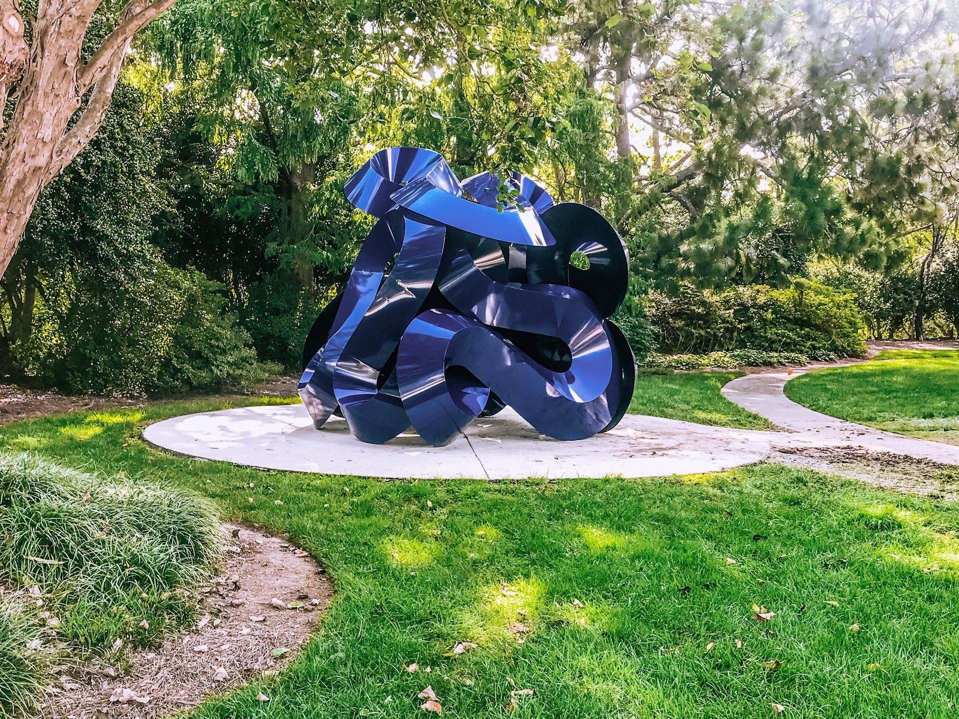 Large, shiny blue abstract sculpture on a white base in a sunny green park.
