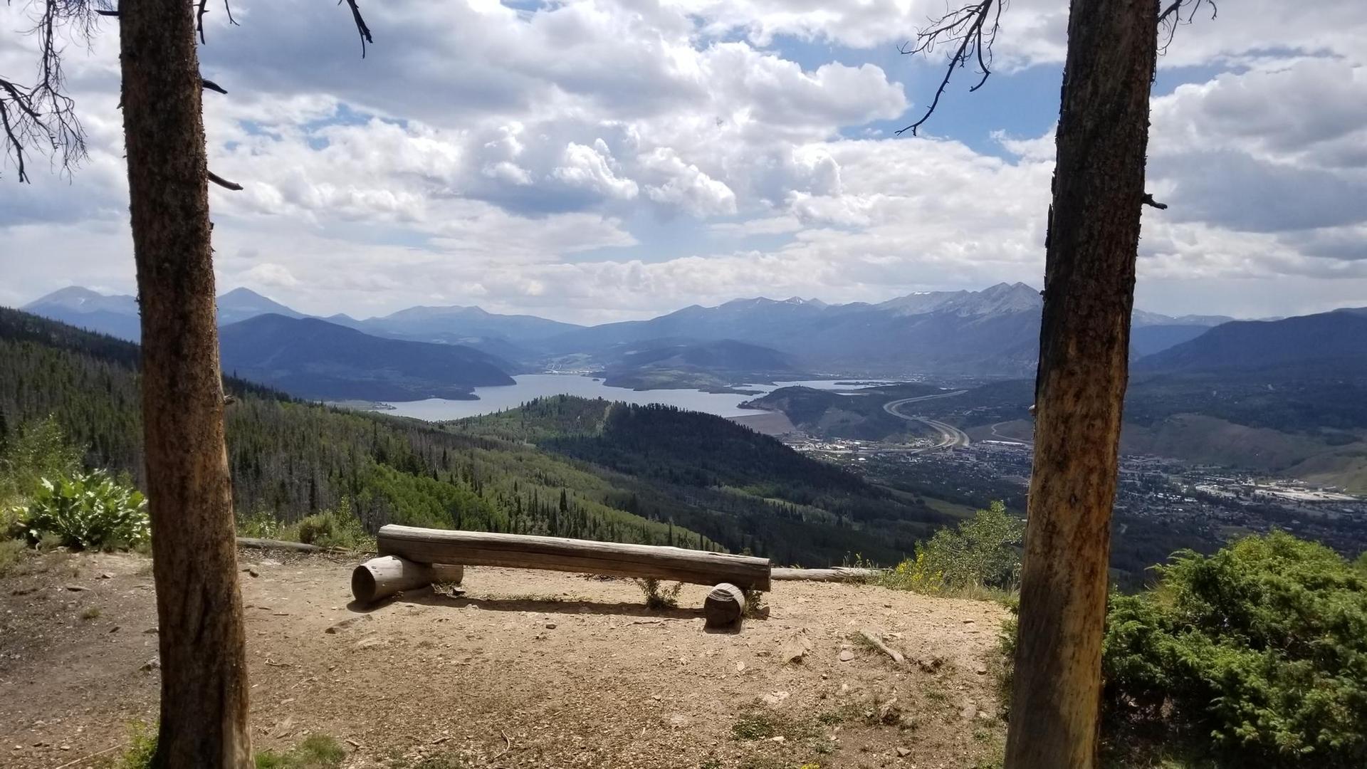 Wooden bench overlooking a mountain lake and valley, framed by trees.
