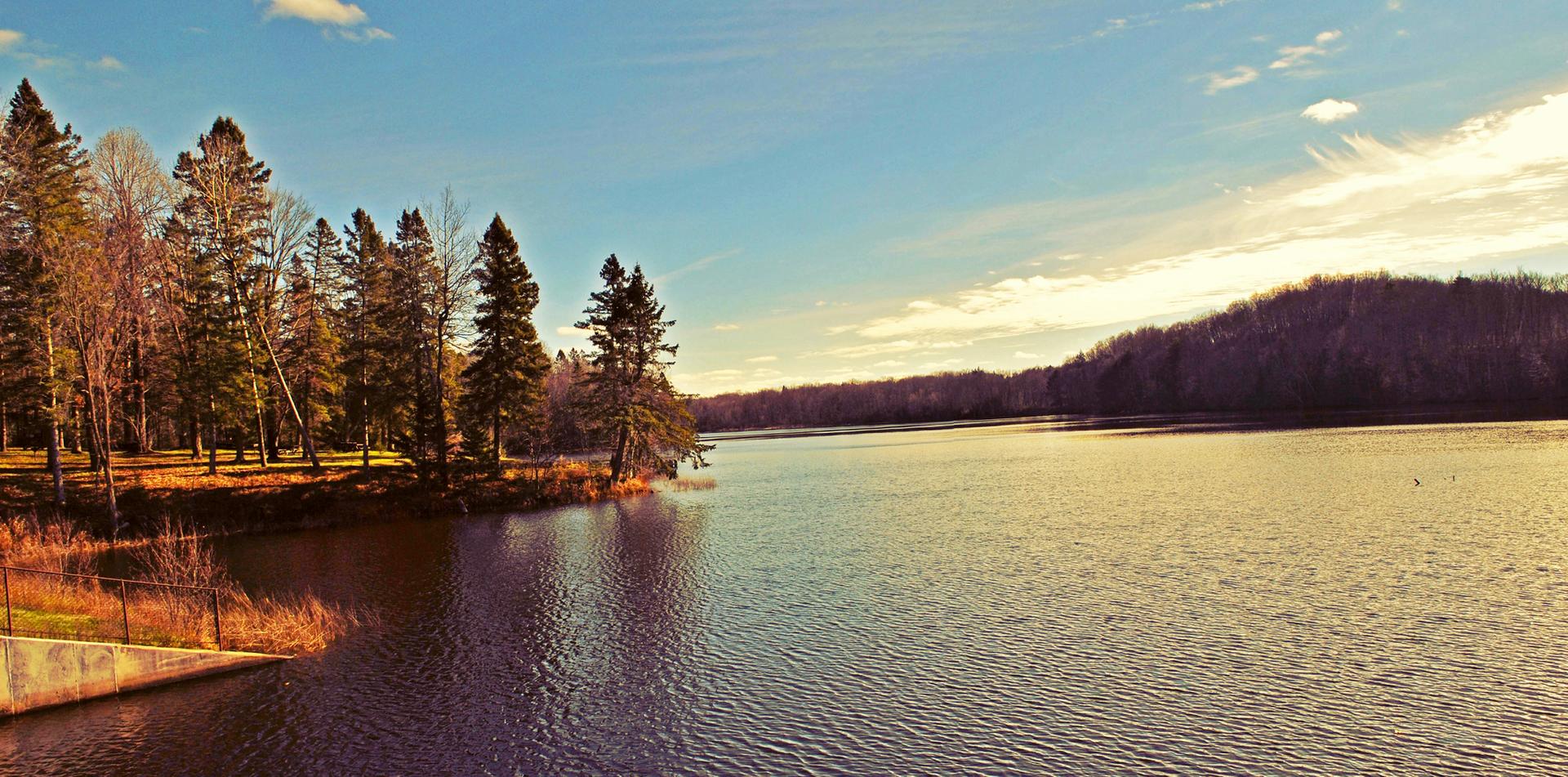 A wide lake with evergreen trees on the left shore under a golden sky.