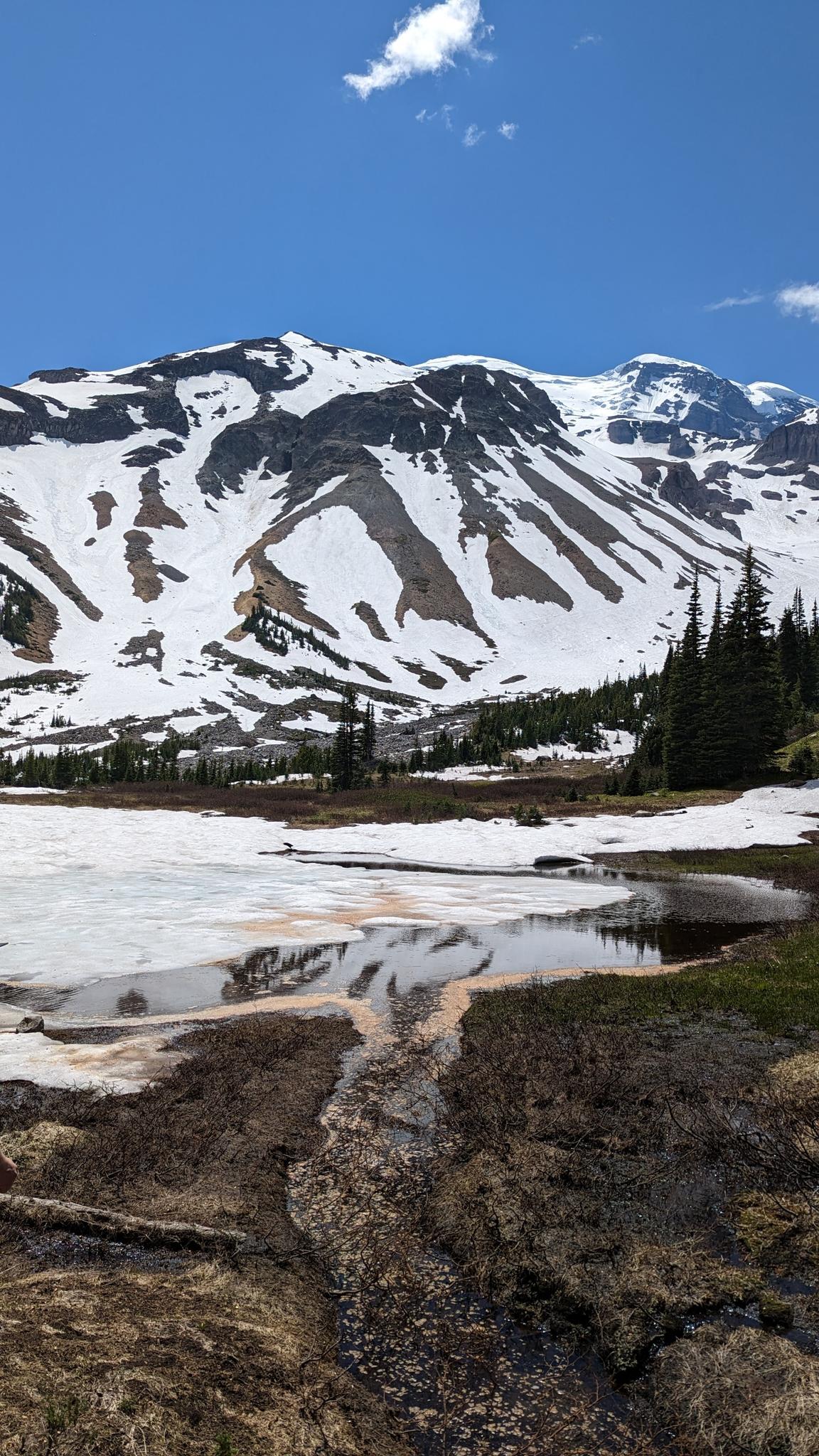 Snow-covered mountain overlooks a partially frozen lake under a blue sky.