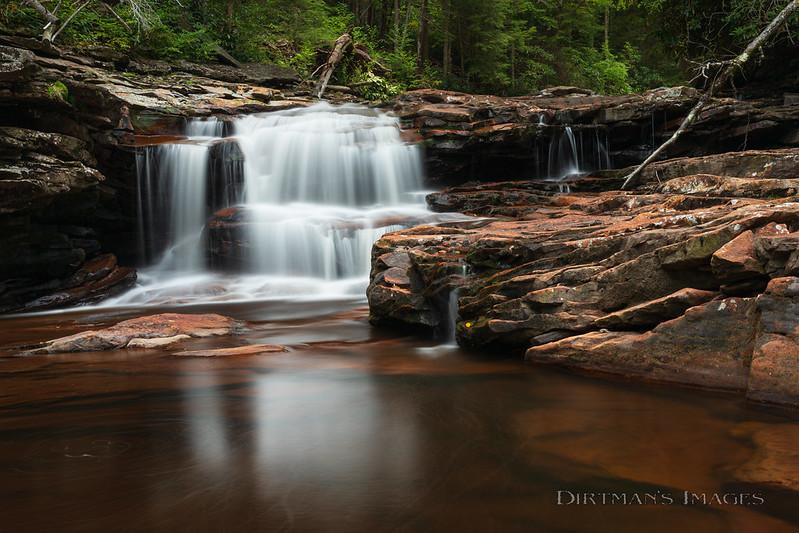 Wide waterfall flowing over red rock ledges into a dark, calm pool.