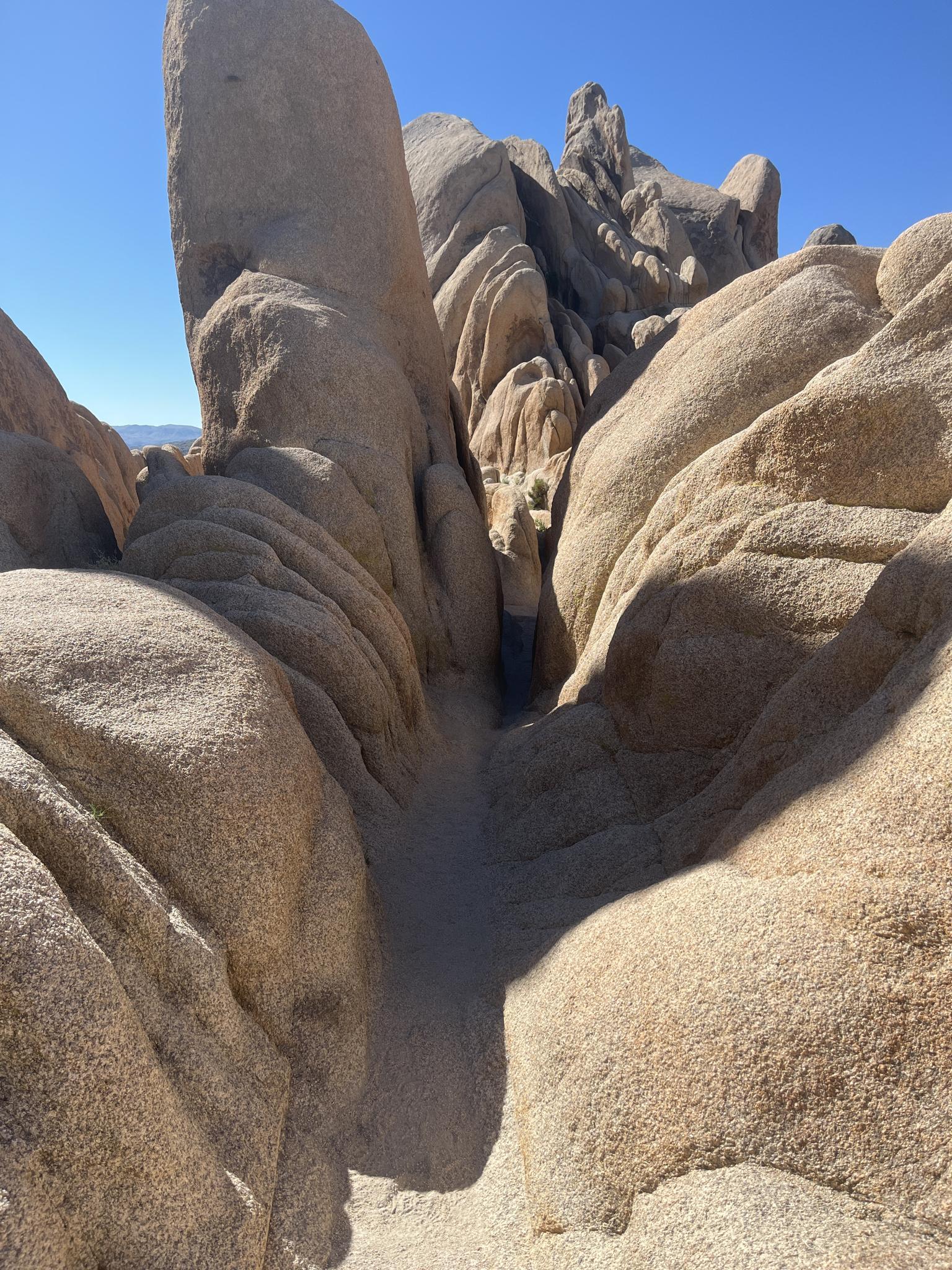 Narrow sandy passage between towering, sunlit rock formations under a clear sky.