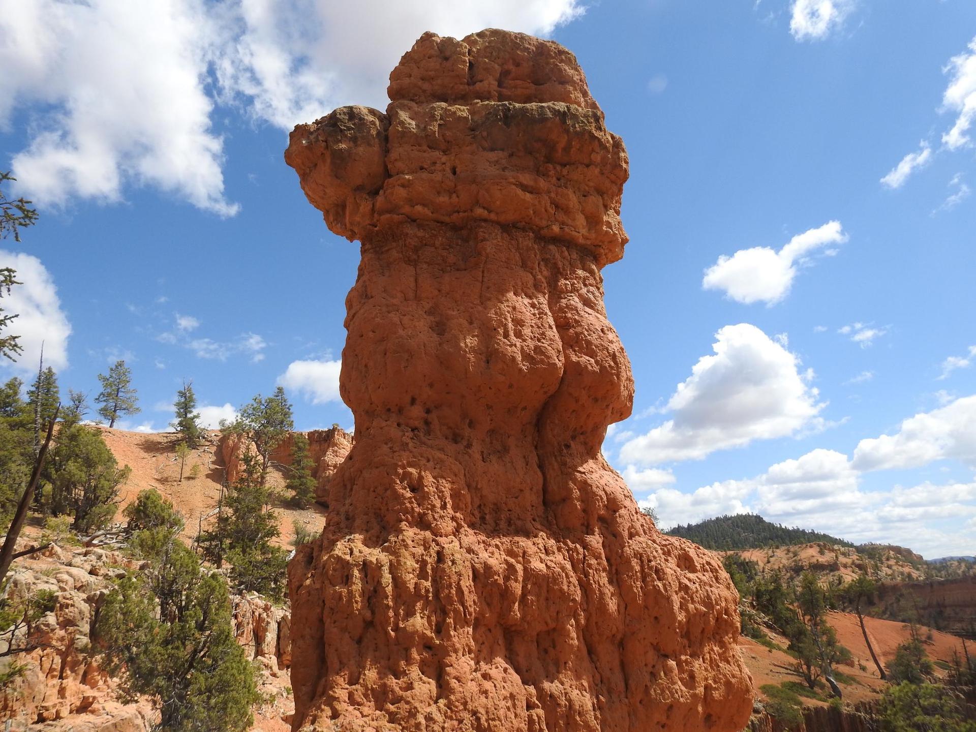 A tall, reddish-orange rock formation against a bright blue sky with white clouds.