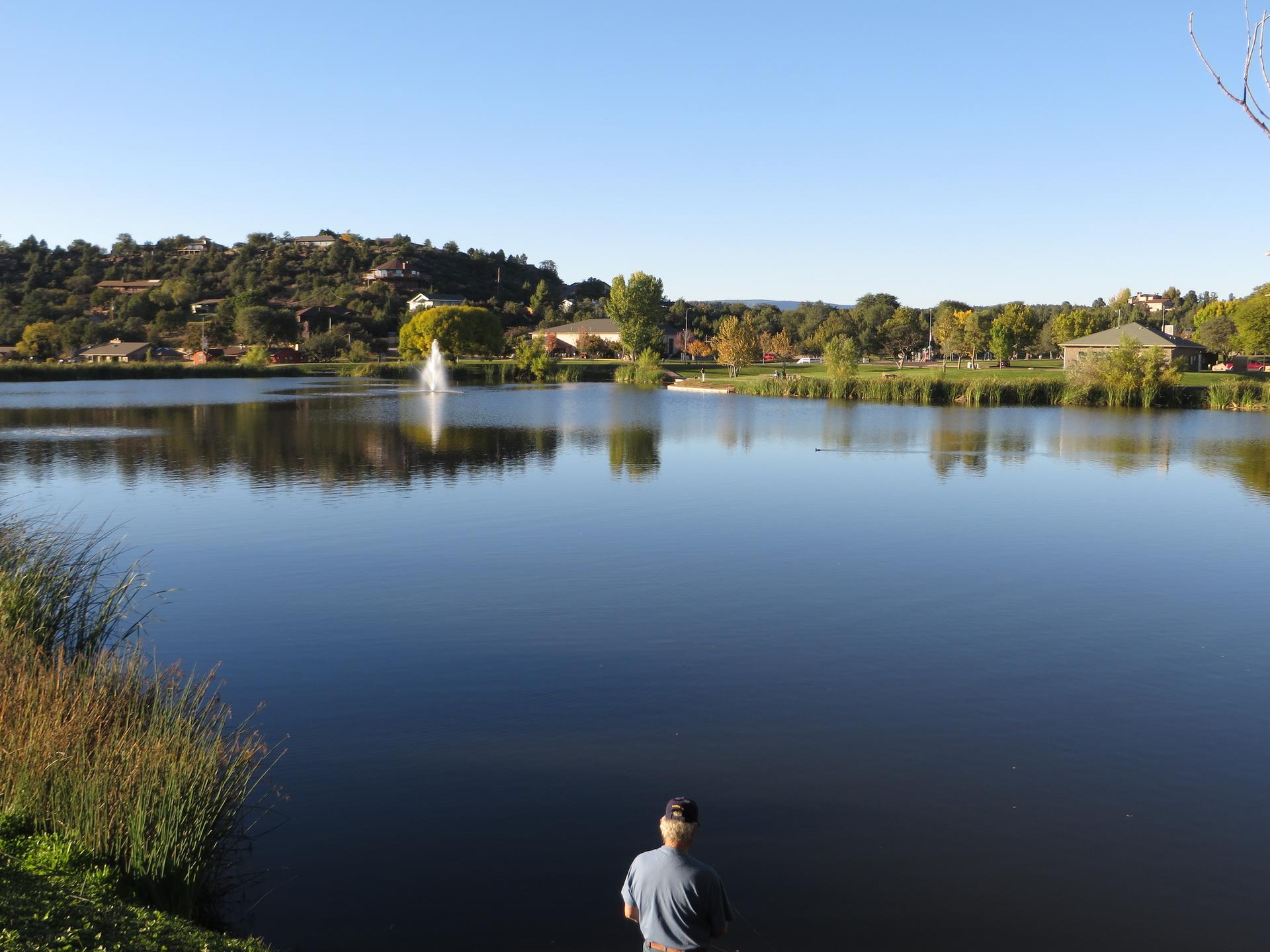 Person fishing by a calm lake with a fountain and distant hills on a clear day.