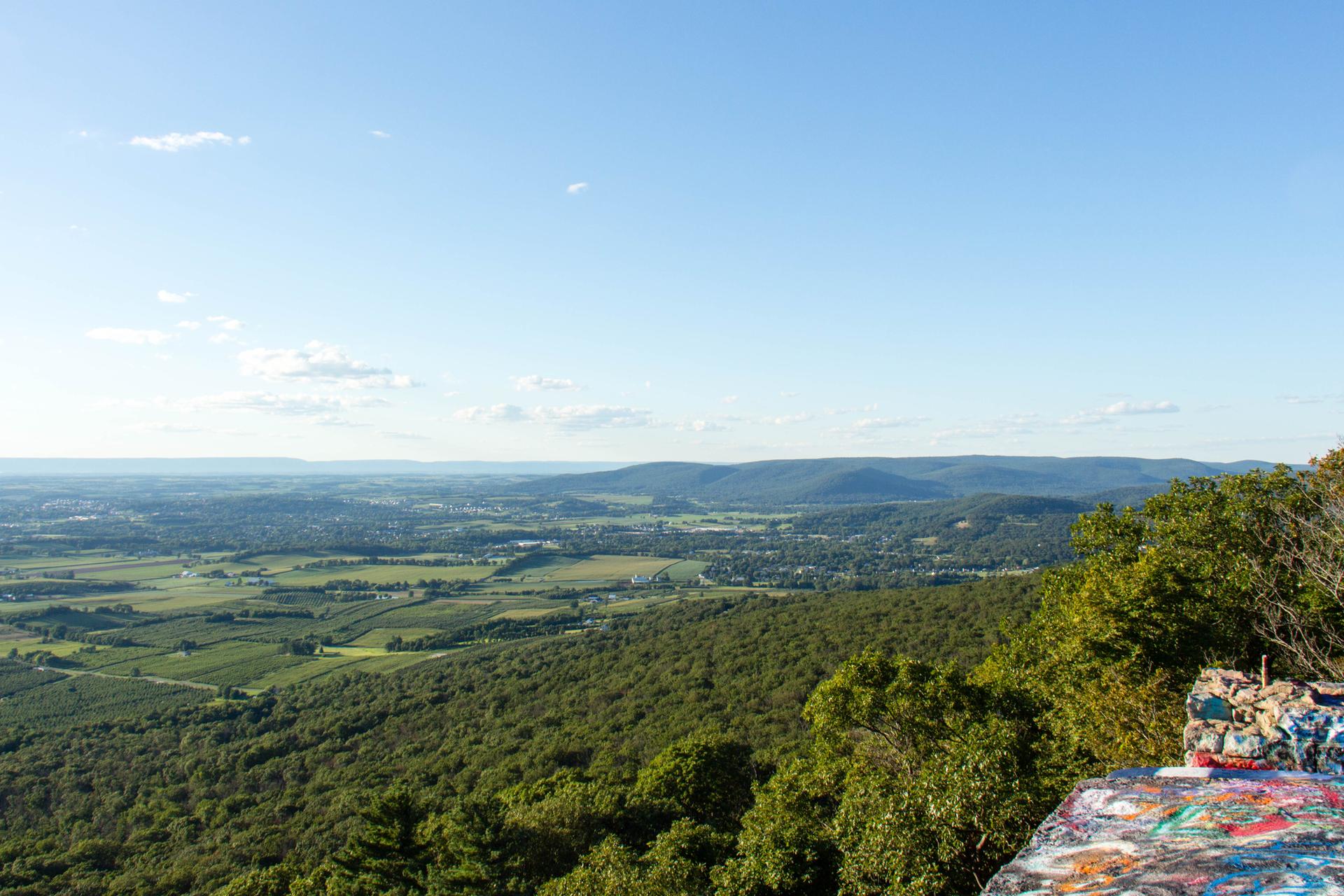 Graffiti-covered rock overlooks a vast green valley under clear skies.