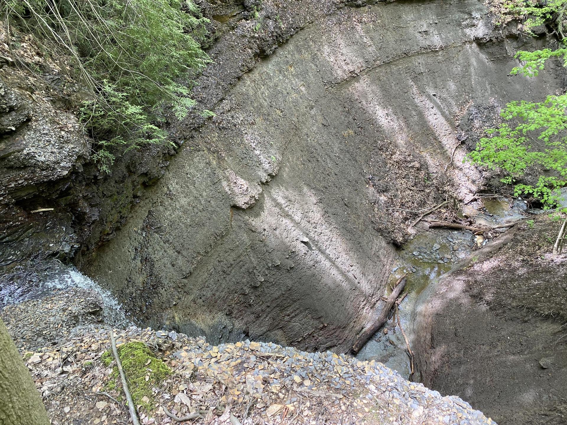 Steep, layered rock formation in a narrow gorge with a stream and dappled sunlight.