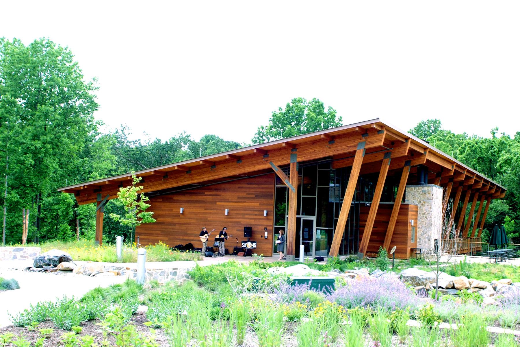 Modern wooden building with musicians playing outside, surrounded by green trees and gardens.
