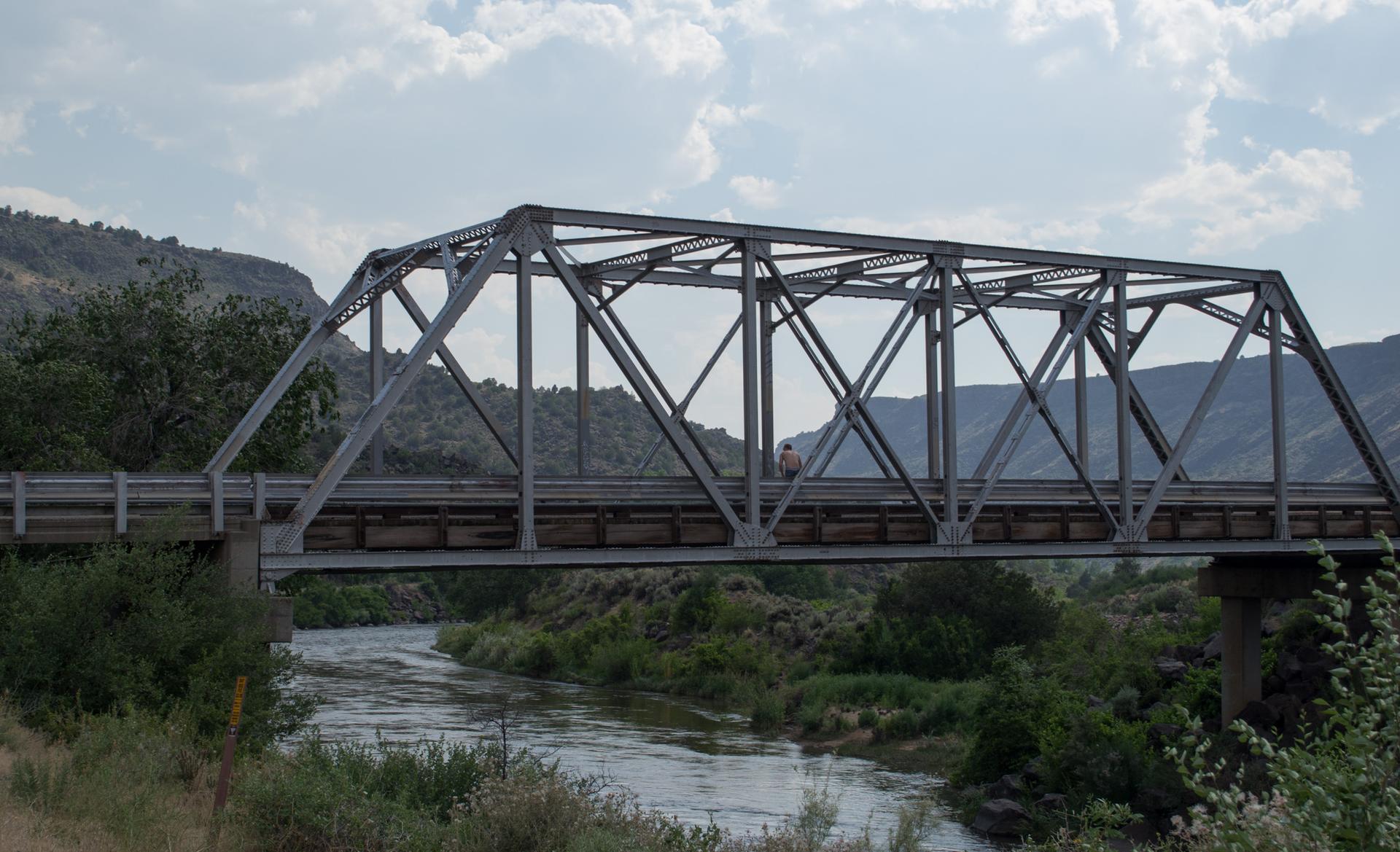 Metal truss bridge spans a river with green banks under a cloudy sky.