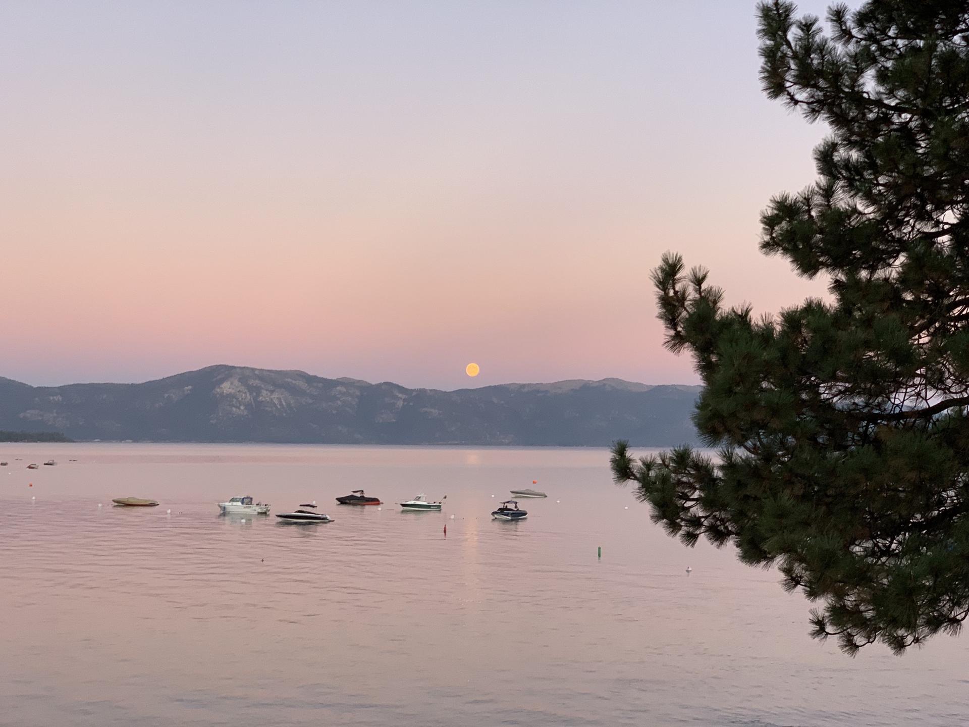 Full moon rising over a lake with boats, mountains, and a pink sky.