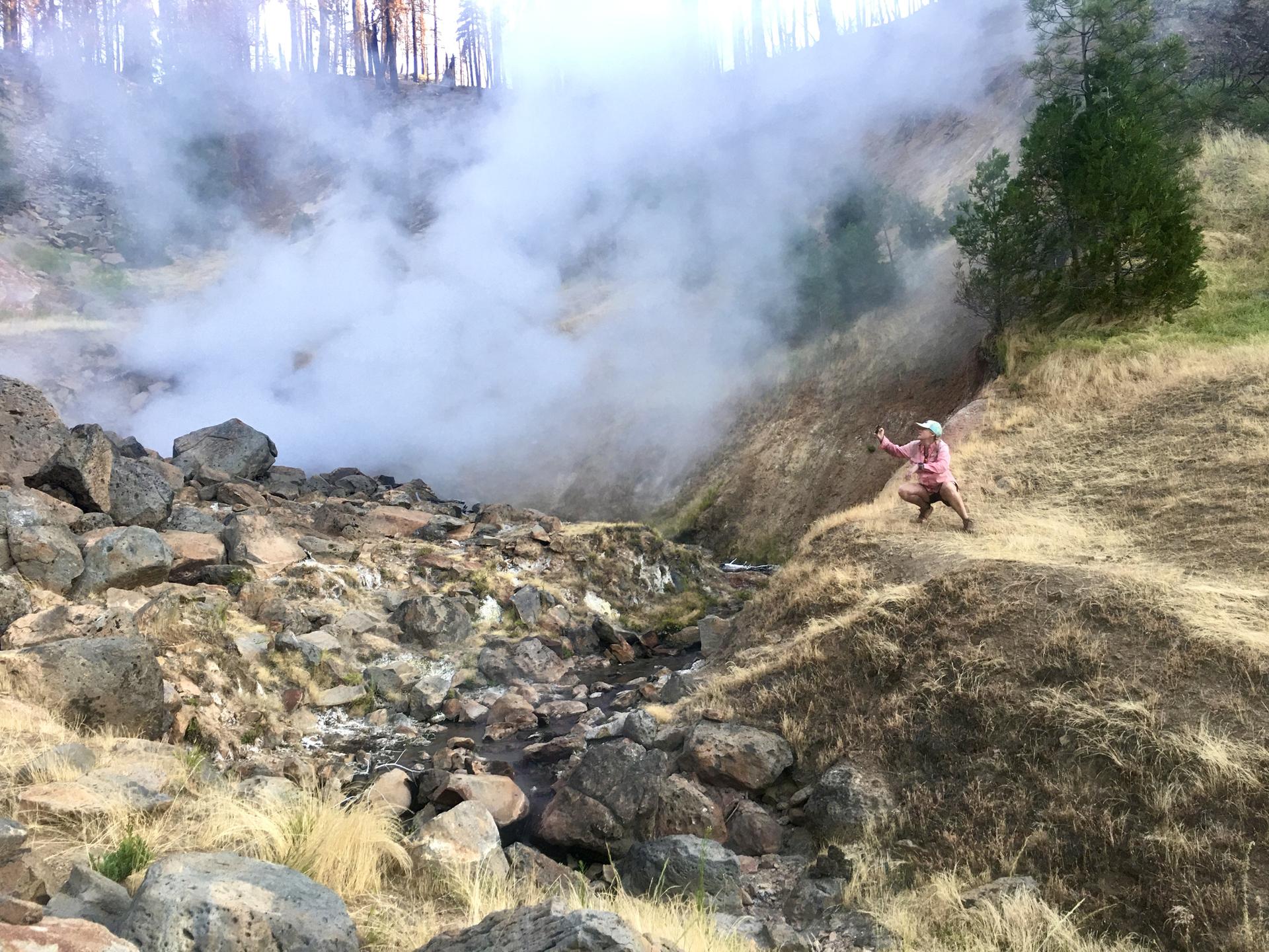 Person crouches near steaming vents in a rocky, grassy, volcanic landscape.