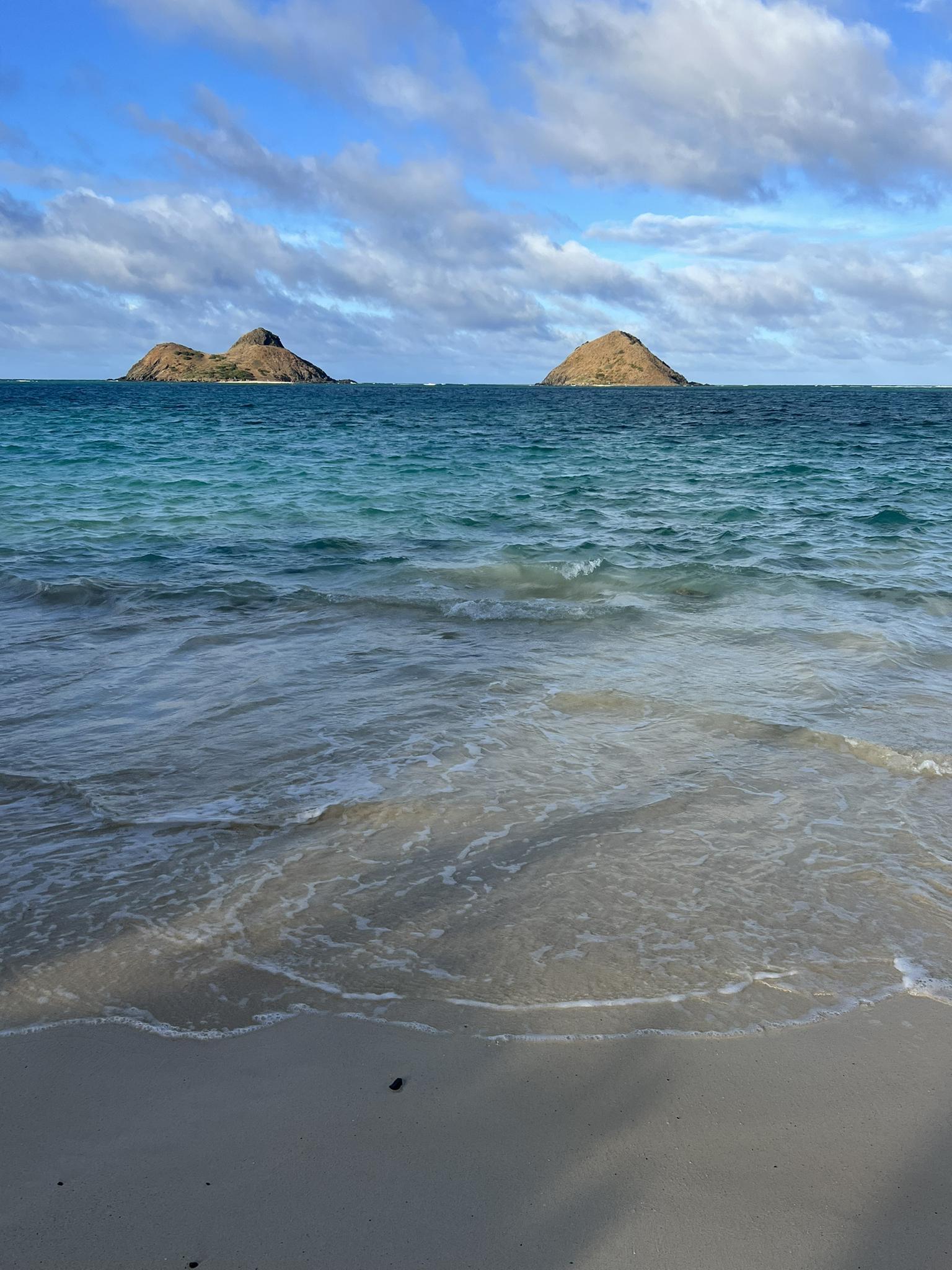 Bright ocean view with two small islands and gentle waves on a sandy beach.