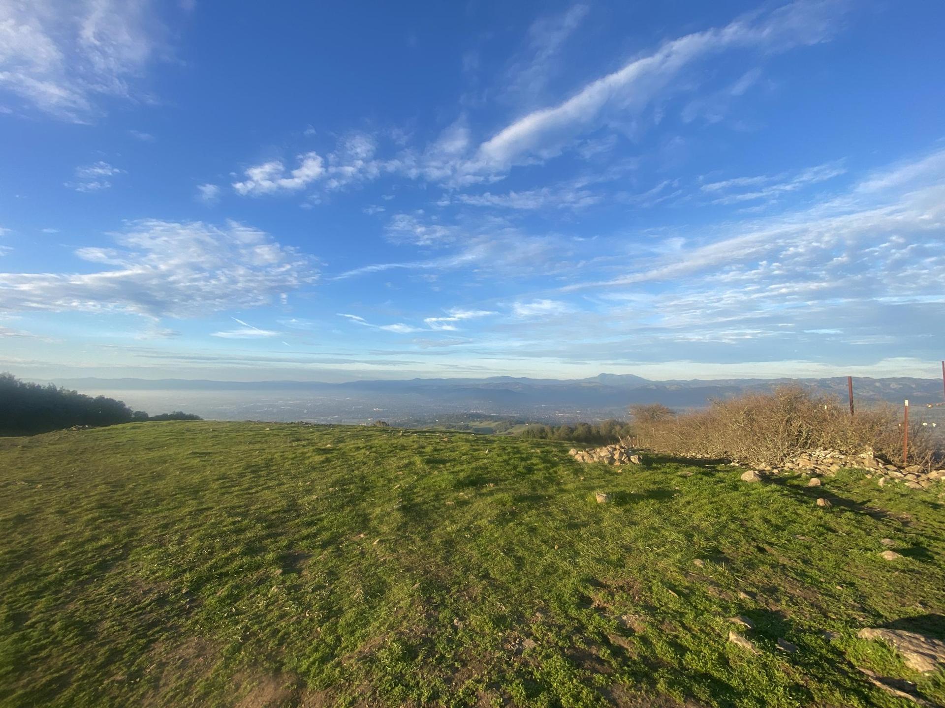 Bright green hillside overlooks a hazy valley under a blue, cloudy sky.