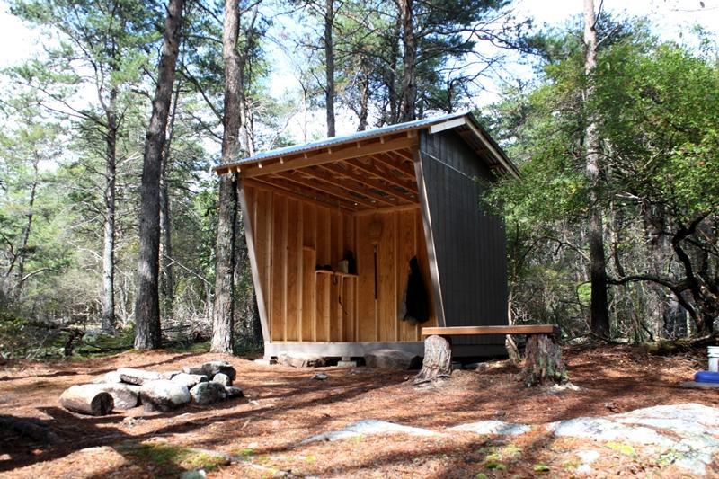 Rustic wooden shelter in a sunlit forest with a fire pit and bench.