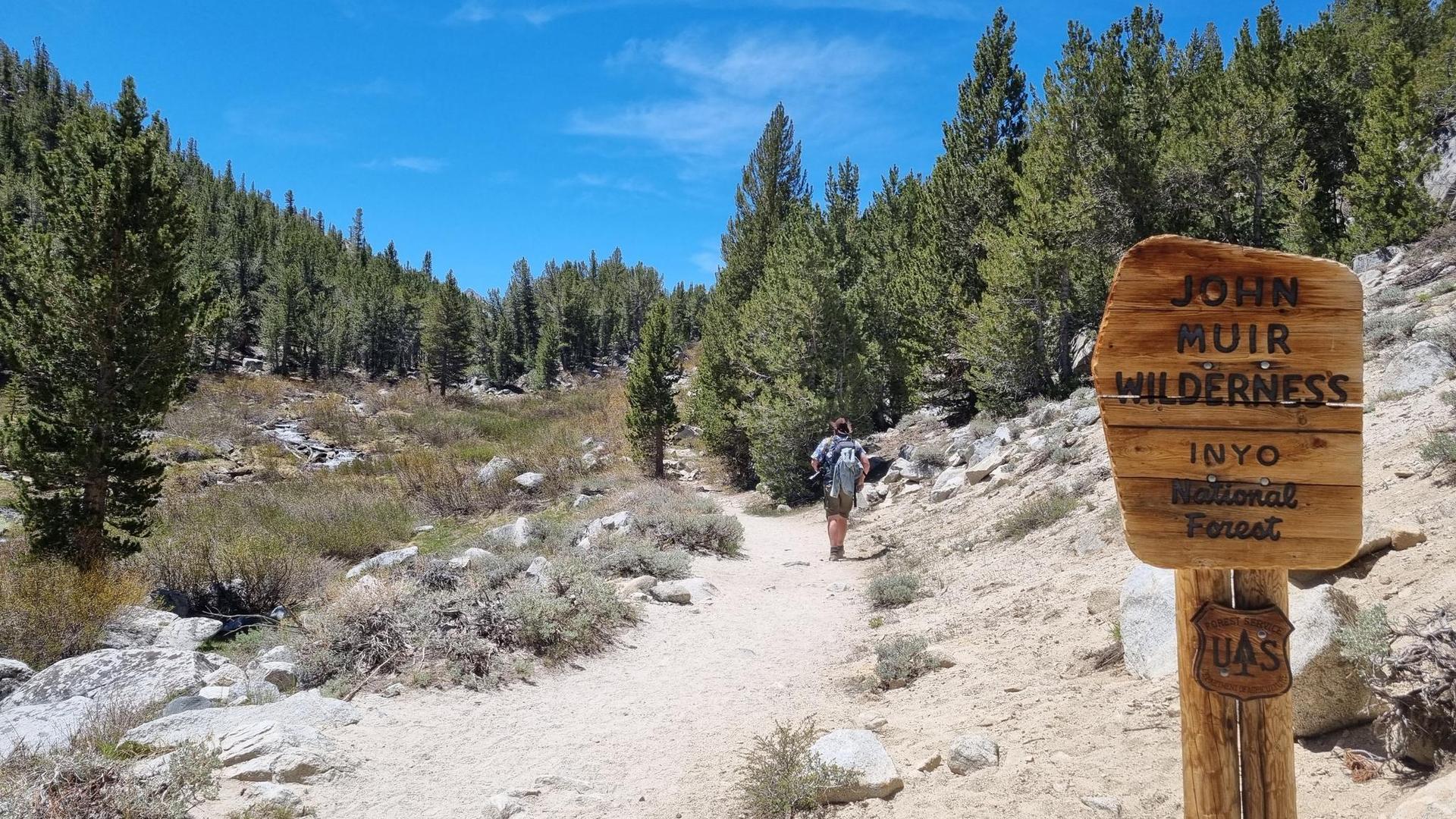A hiker walks a sunny dirt path through a forested mountain landscape with a wooden sign.