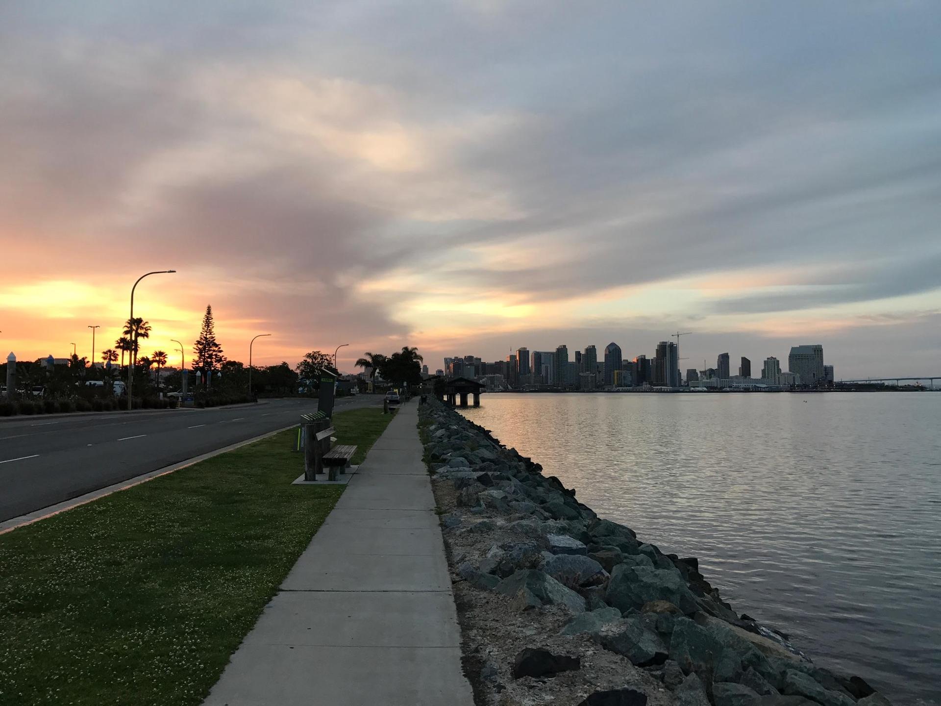 Waterfront path and road at sunset with a distant city skyline across the water.