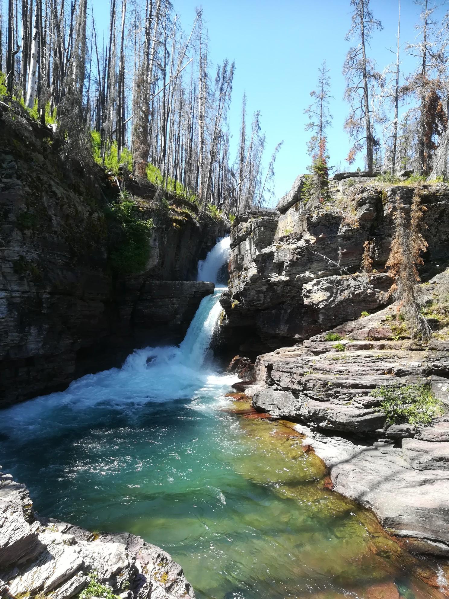 Waterfall cascading into a turquoise pool, surrounded by rocky cliffs and dead trees.