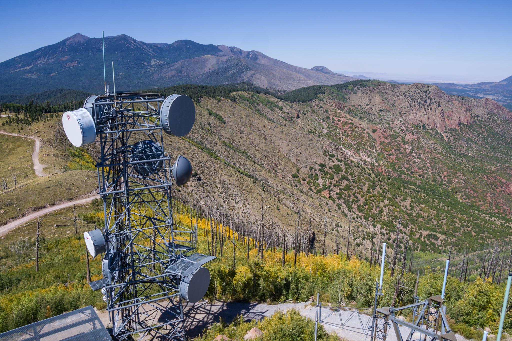 A large communication tower stands on a mountain with distant peaks under clear sky.