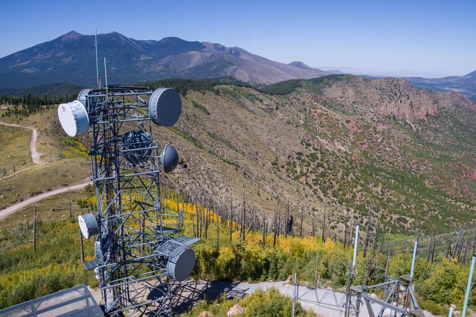 A large communication tower stands on a mountain with distant peaks under clear sky.