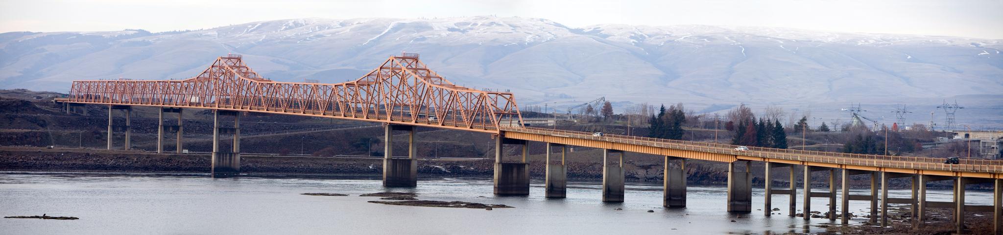 A long orange truss bridge spans a river, with snow-capped mountains beyond.