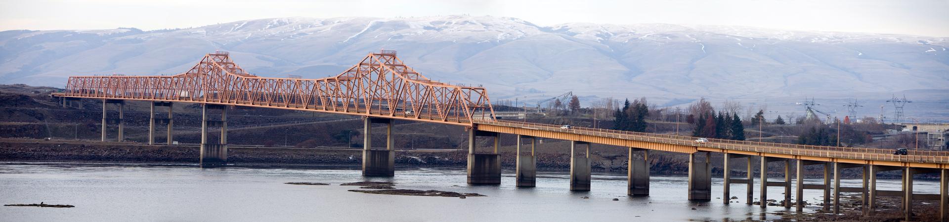 A long orange truss bridge spans a river, with snow-capped mountains beyond.