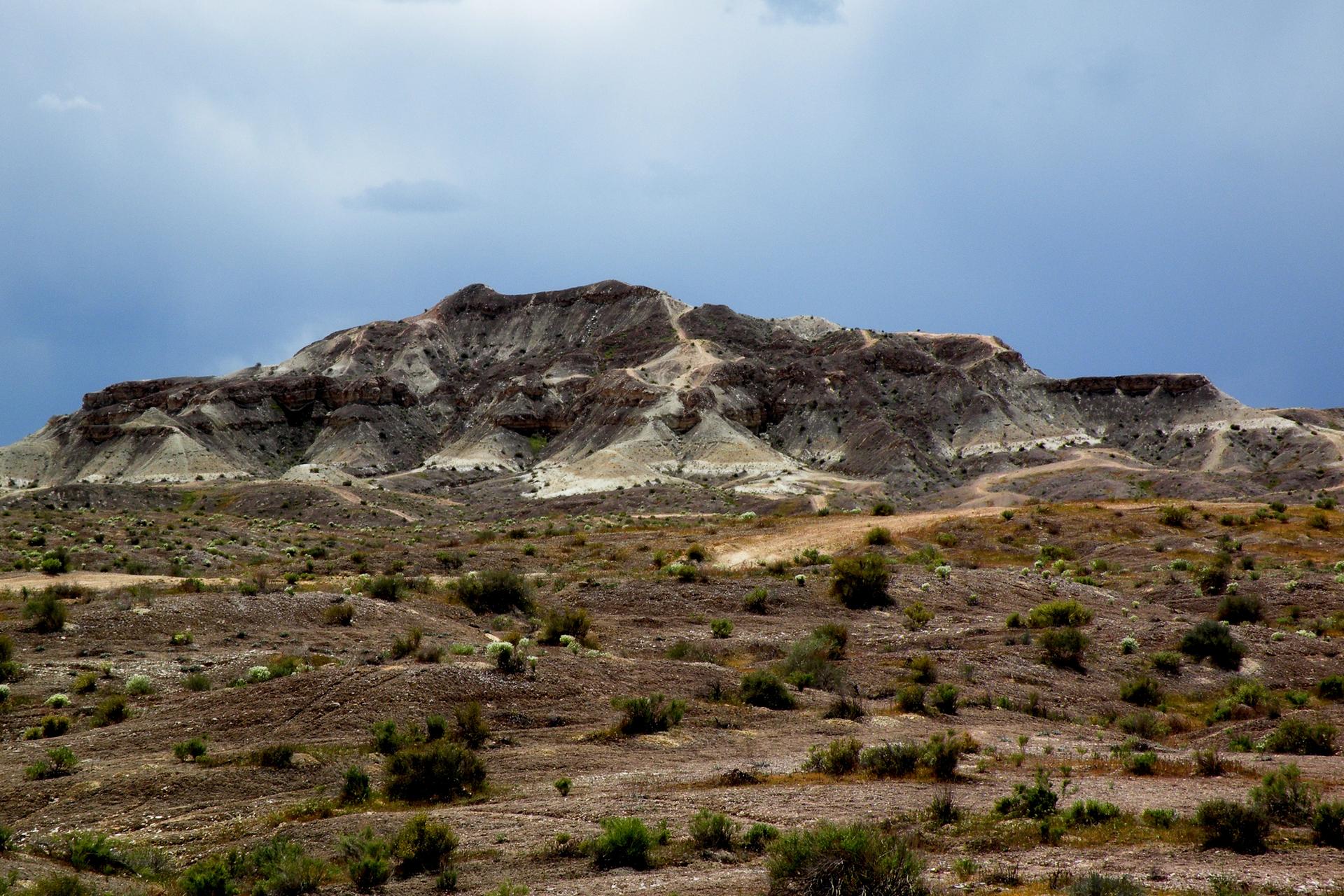 Rocky, layered mountain dominates an arid landscape under a dramatic cloudy sky.