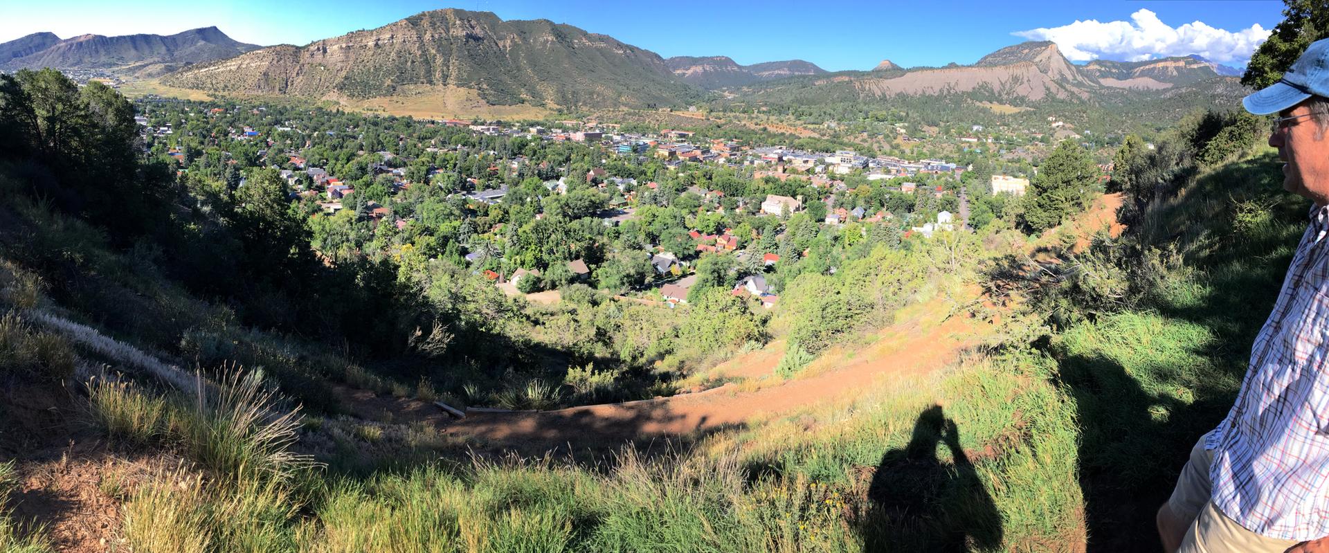 A person overlooks a sunny town in a green valley with distant mountains.