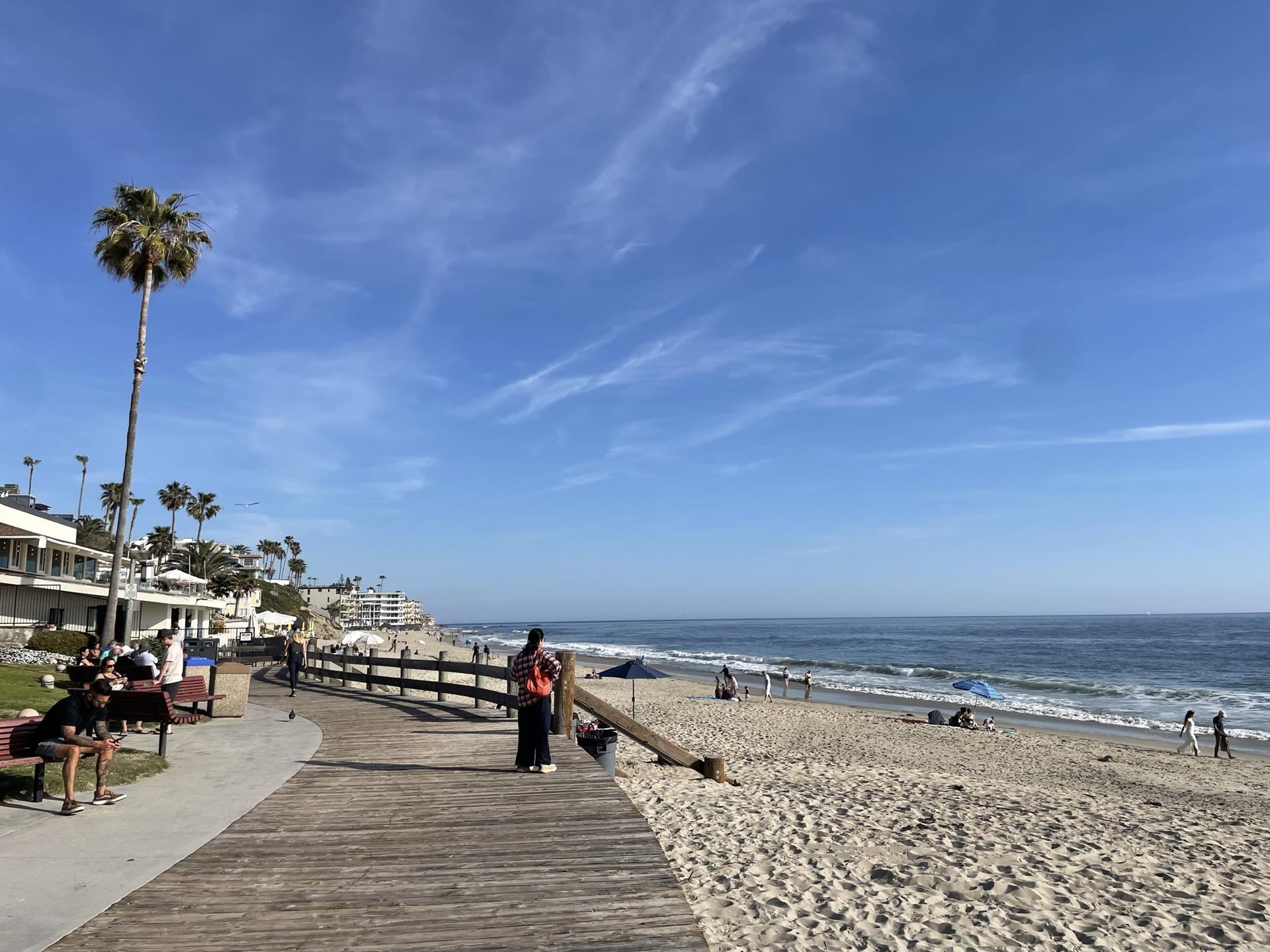 Sunny beach with boardwalk, palm trees, ocean, and people relaxing.