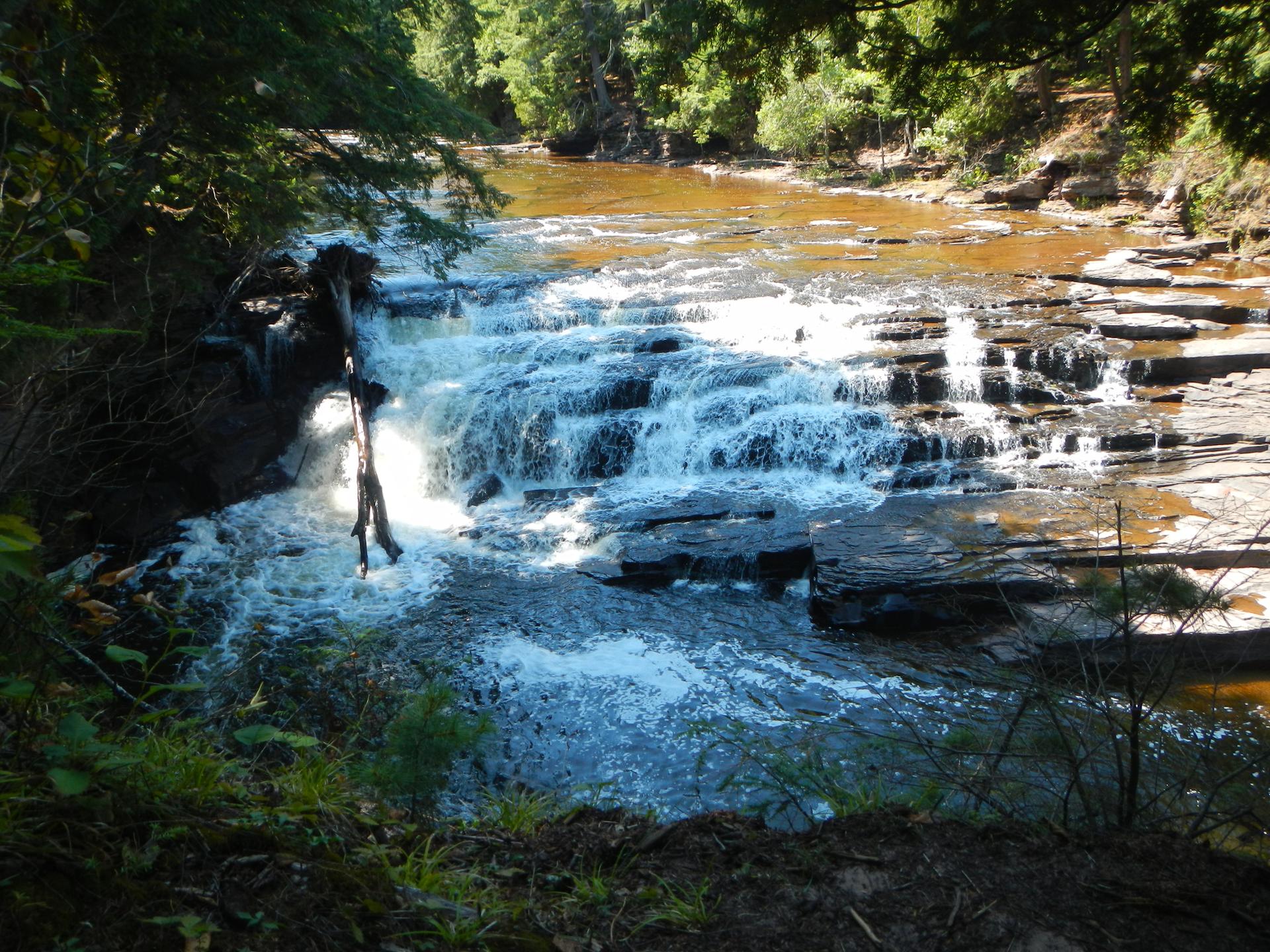Sunlight illuminates a waterfall flowing over dark, stepped rocks in a forest.
