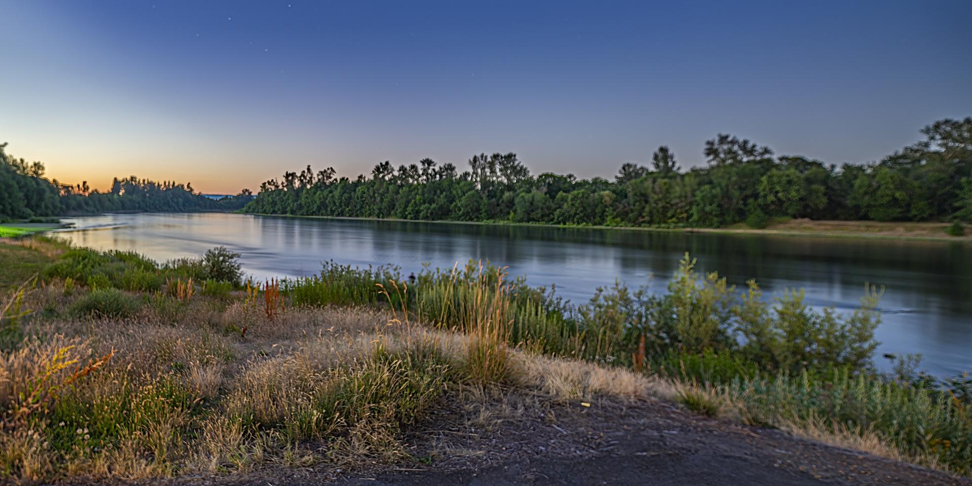 Wide river flowing past tree-lined banks at dusk under a fading sky.