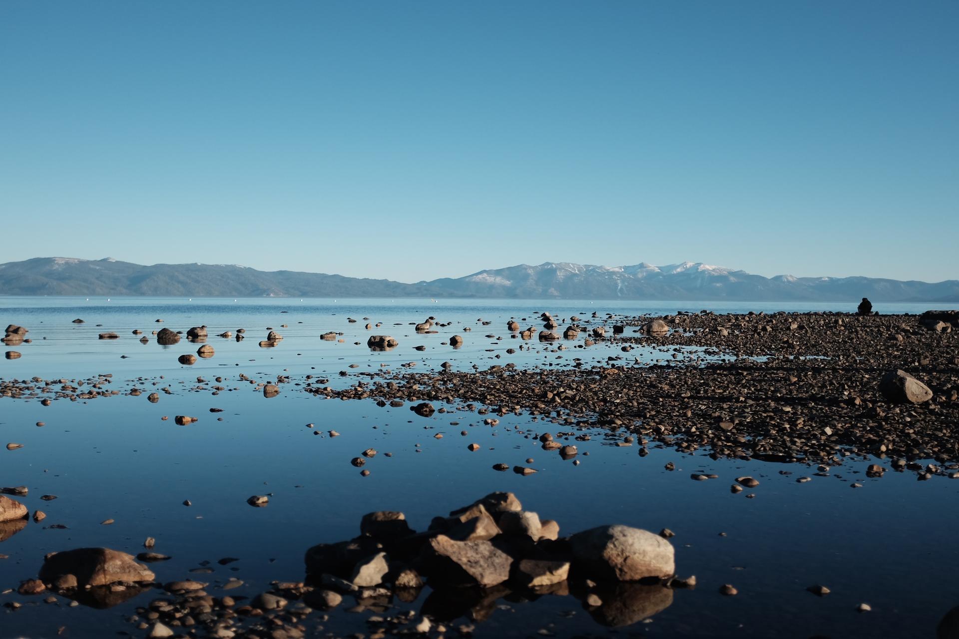 Rocky shoreline and calm lake under a clear sky with distant mountains.