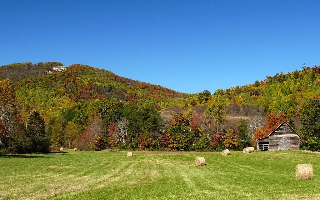 A rustic barn and hay bales in a sunny field with autumn mountains.