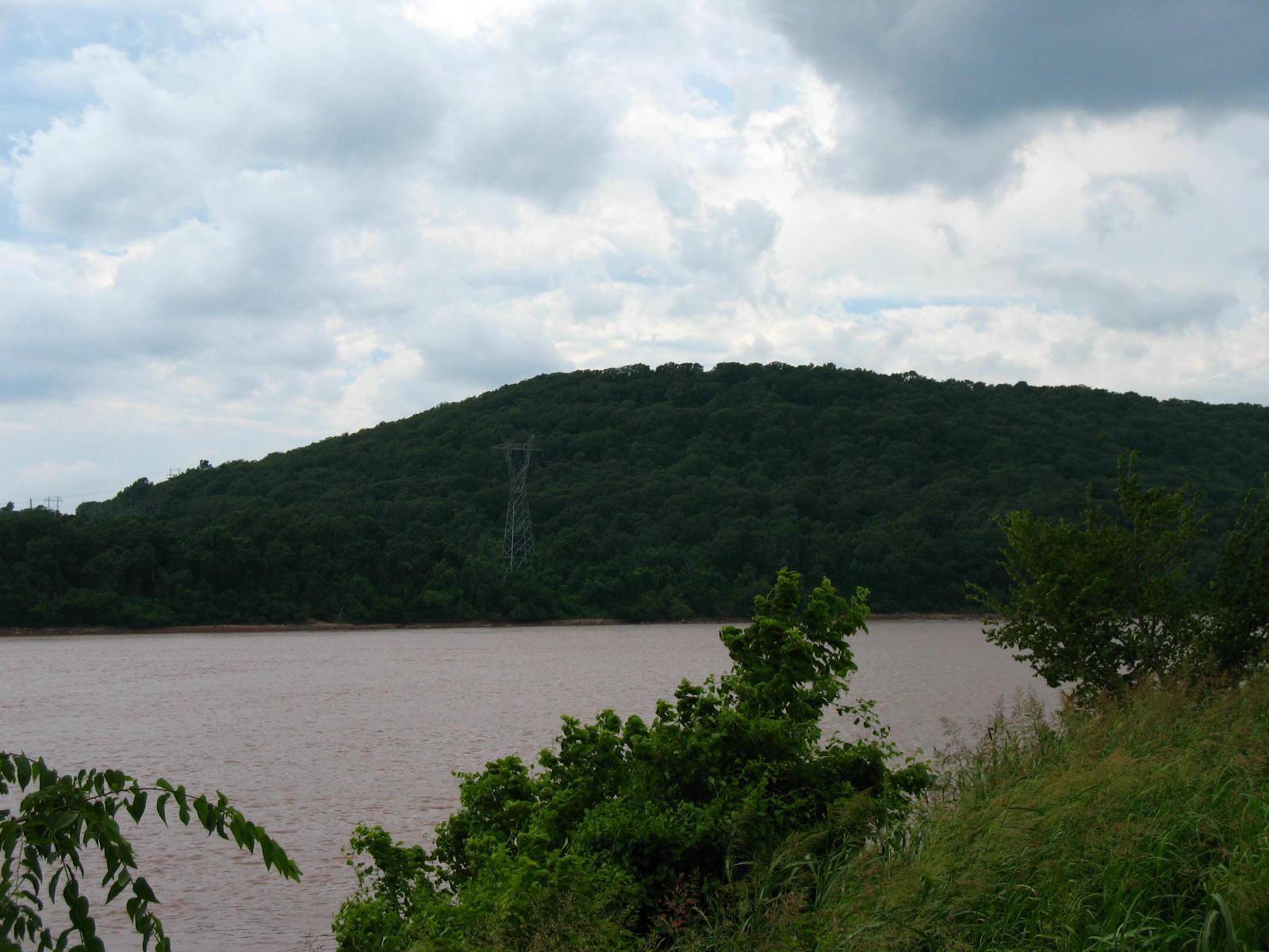 Wide river with a densely forested hill under a cloudy sky.