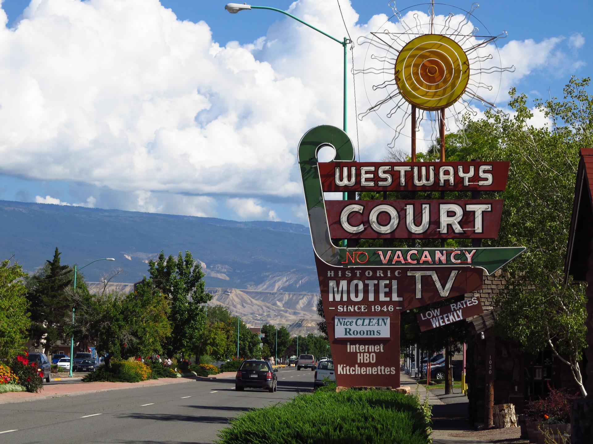 A vintage motel sign with a sun graphic stands by a road with mountains.