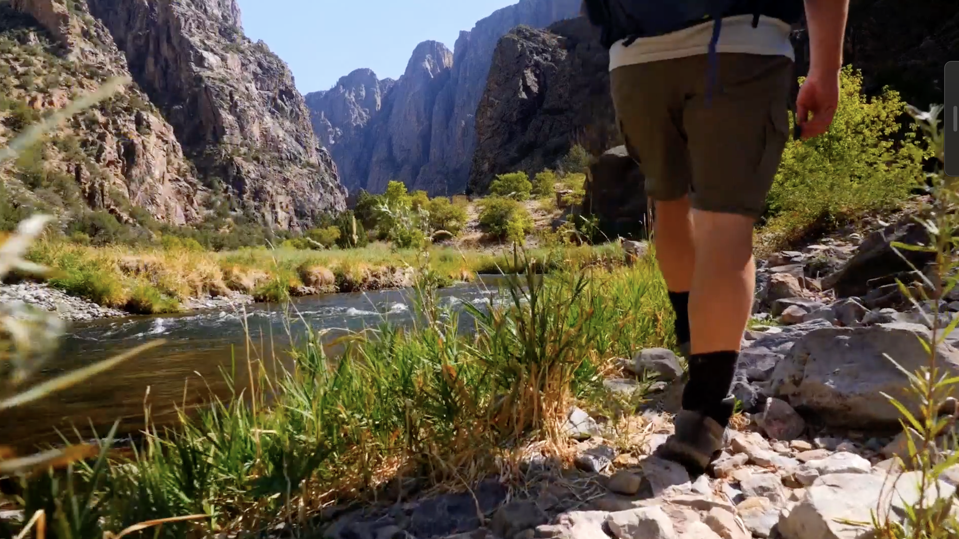 Hiker walks along a rocky riverbank with tall canyon walls in daylight.