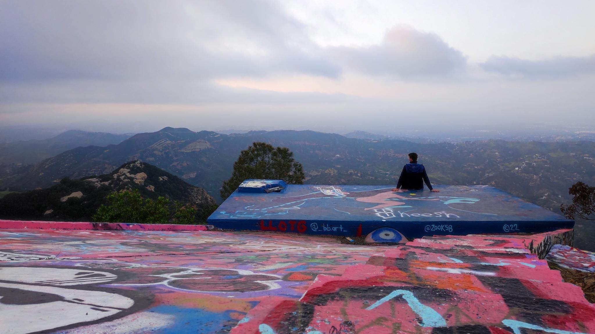 Person sits on a graffiti-covered platform overlooking cloudy mountains. - Backbone Trail Loop